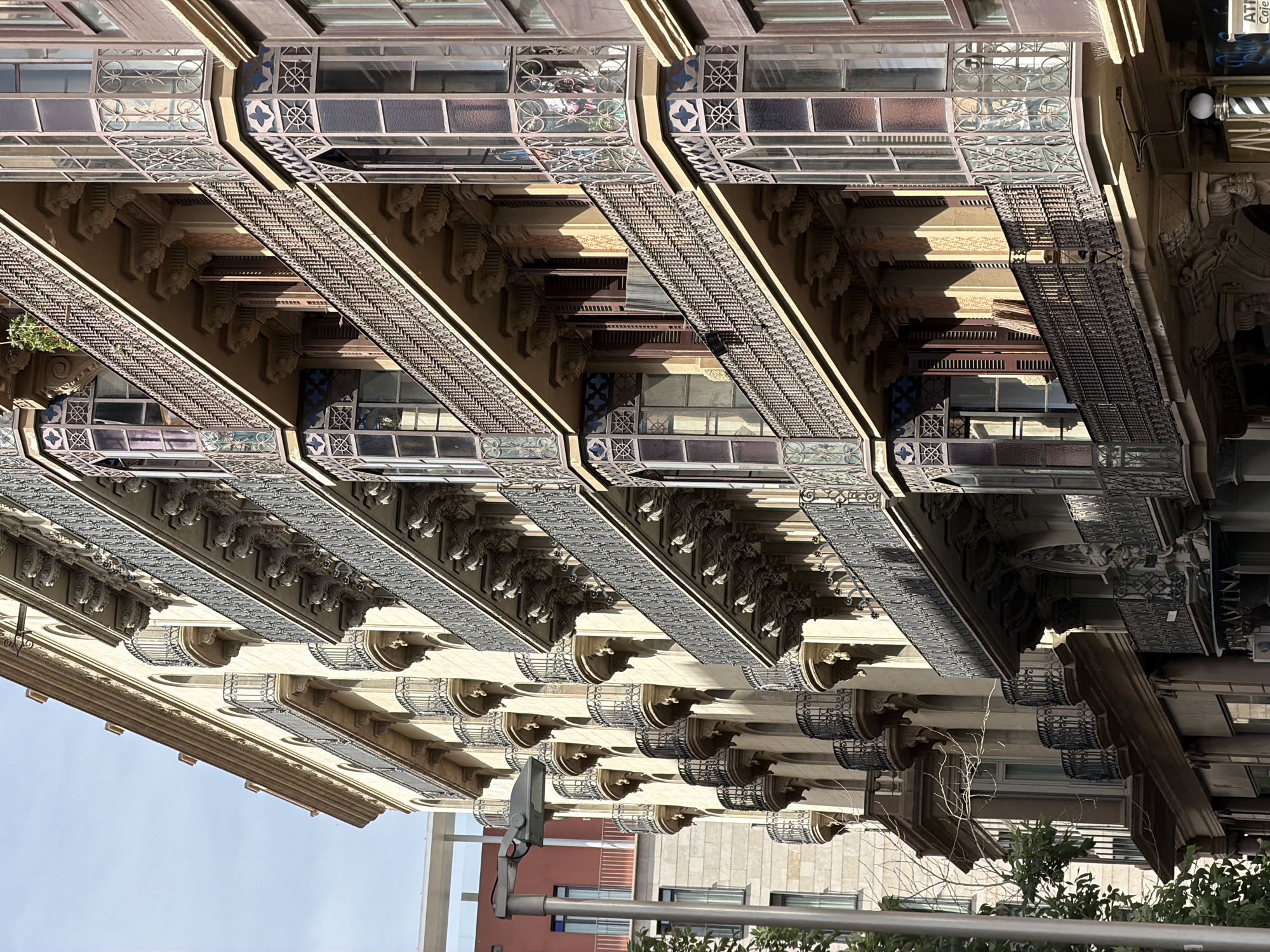 This is a street-level photograph of a historic building facade in Barcelona, Spain, specifically on the Passeig de Gràcia. The building features multiple stories with ornate balconies supported by decorative brackets. The balconies have intricate wrought-iron railings and are adorned with stained glass elements in the bay windows. Sunlight casts strong shadows across the facade, highlighting the architectural details. In the foreground, a street lamp and a portion of a tree are visible. The building to the left of the main subject is a more modern structure with a red facade and a white horizontal element. The overall scene depicts a typical urban streetscape of Barcelona with its characteristic modernist architecture.