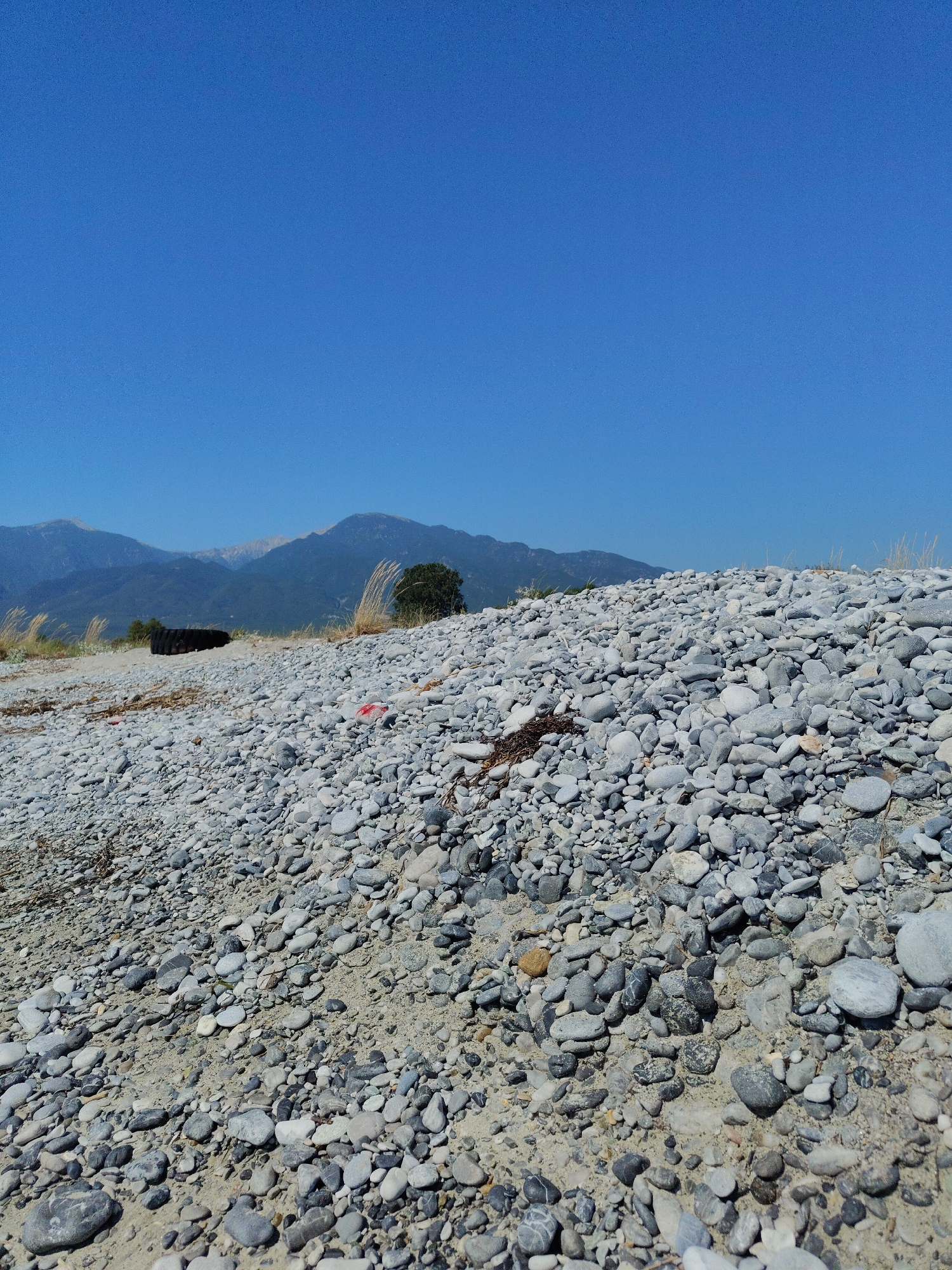 The image depicts a sunlit, outdoor scene with a clear, bright blue sky dominating the upper portion of the frame. The foreground is filled with a dense layer of light-colored pebbles and sand, with some larger stones interspersed. A black tire is visible in the middle ground, partially obscured by the pebbles. Behind the tire, a dark green tree stands out against the landscape. In the background, a range of mountains stretches across the horizon, with the peaks of some mountains showing hints of lighter coloration, possibly due to snow or rock. The terrain appears to be a beach or a dry riverbed, given the abundance of stones and the arid vegetation. The overall impression is one of a natural, undeveloped area.