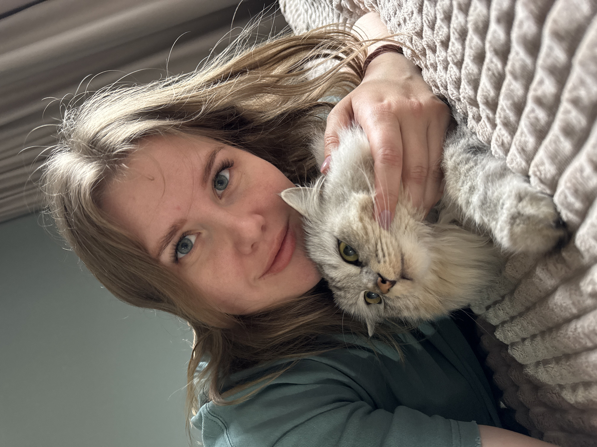 An indoor, close-up portrait shows a young adult woman with fair skin, light blue eyes, and long, wavy blonde hair, smiling subtly while holding a fluffy grey and beige cat. The woman is positioned on the left side of the frame, looking directly at the viewer, with her head tilted. She is wearing a teal or green hooded top. Her right hand is gently holding the cat close to her face, its head resting near her chin. The cat, with striking golden-green eyes, appears calm and content, looking towards the viewer with its head slightly tilted, mimicking the woman's posture.
The background consists of a plain, light grey-green wall on the left, and a textured, light beige or cream blanket with a woven pattern on the right, which the woman's arm is partially resting on. The image has a warm and intimate feel, capturing a moment of affection between the woman and her pet. The lighting is soft and diffused, characteristic of indoor natural or ambient light. There is no visible text in the image. The scene conveys a sense of comfort and companionship, set in Gornyi (Dvurechenskoe S/P), Russia.