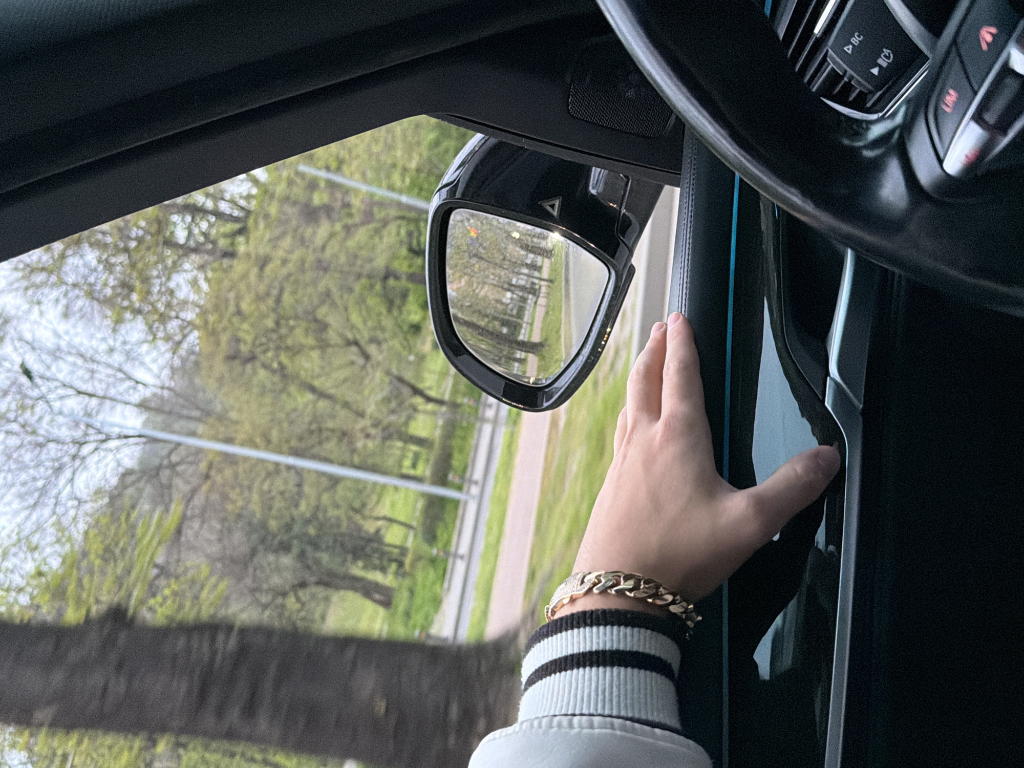 An eye-level shot captures a person's right hand and forearm resting on the dark interior door panel of a car. The hand, adorned with a chunky gold curb-link bracelet featuring a diamond-encrusted link, wears a white sleeve cuff with two black stripes.
Beyond the car's window, lush green trees and foliage are visible, suggesting a vibrant outdoor environment, likely during spring or summer. The scene is bright, indicating daytime. A side-view mirror of the car is prominent, reflecting a tree-lined road or pathway with a light pole in the distance. The mirror itself shows a small triangular indicator, possibly for a blind-spot monitoring system.
Part of the car's black leather-wrapped steering wheel and control buttons are visible in the upper right. Buttons feature symbols such as "BC", play/pause, a phone icon, and "LIM", suggesting modern vehicle technology. The overall setting appears to be a drive or a parked moment in an area surrounded by nature. The location is Unknown.