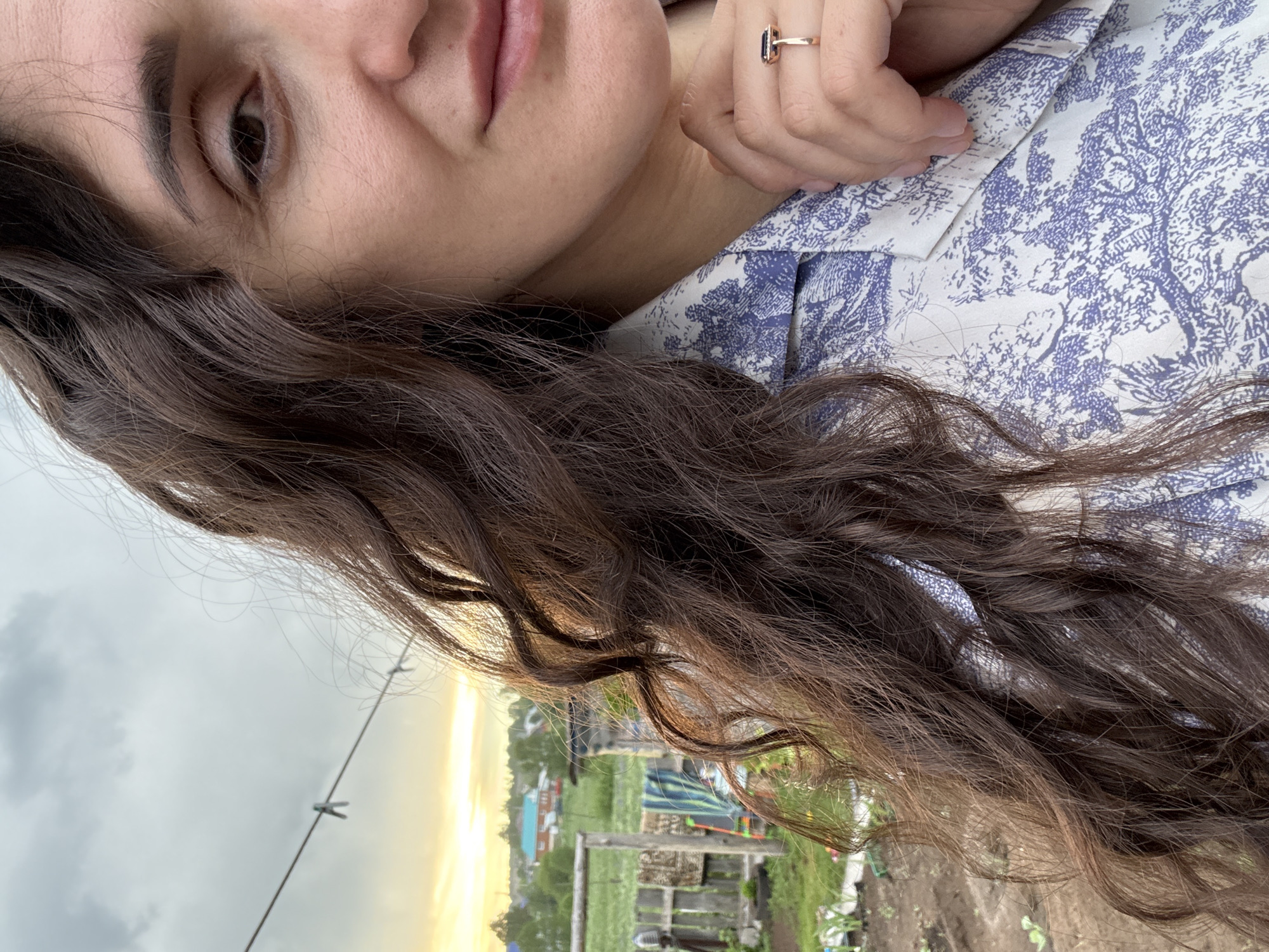 A close-up, slightly off-center, and rotated image shows a woman in Уральское, Russia. Her face is partially visible, with a focus on her eye, nose, and mouth. She has long, dark curly hair. She wears a gold ring with a dark blue stone. The woman's left hand rests near her collar, which is patterned with a blue and white floral design. The background reveals a glimpse of the outdoors, with a view of houses, greenery, and the sky. A clothesline with clothespins is visible. The sky appears overcast with a golden sunset.