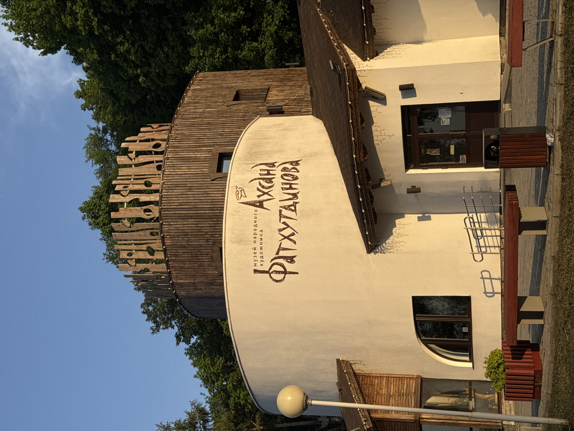 The image shows the exterior of the Museum of Folk Artist Fatkhutdinova in Krasny Klyuch, Russia. The building has a unique architectural style, featuring a rounded tower-like structure with wooden planking details at the top. A sign on the side of the tower displays the name of the museum in a decorative font. Part of the building has a white wall that connects to a rectangular building. The exterior includes windows, doors, a bench, bicycle rack, and decorative lighting. Green trees and a clear blue sky form the backdrop of the scene.