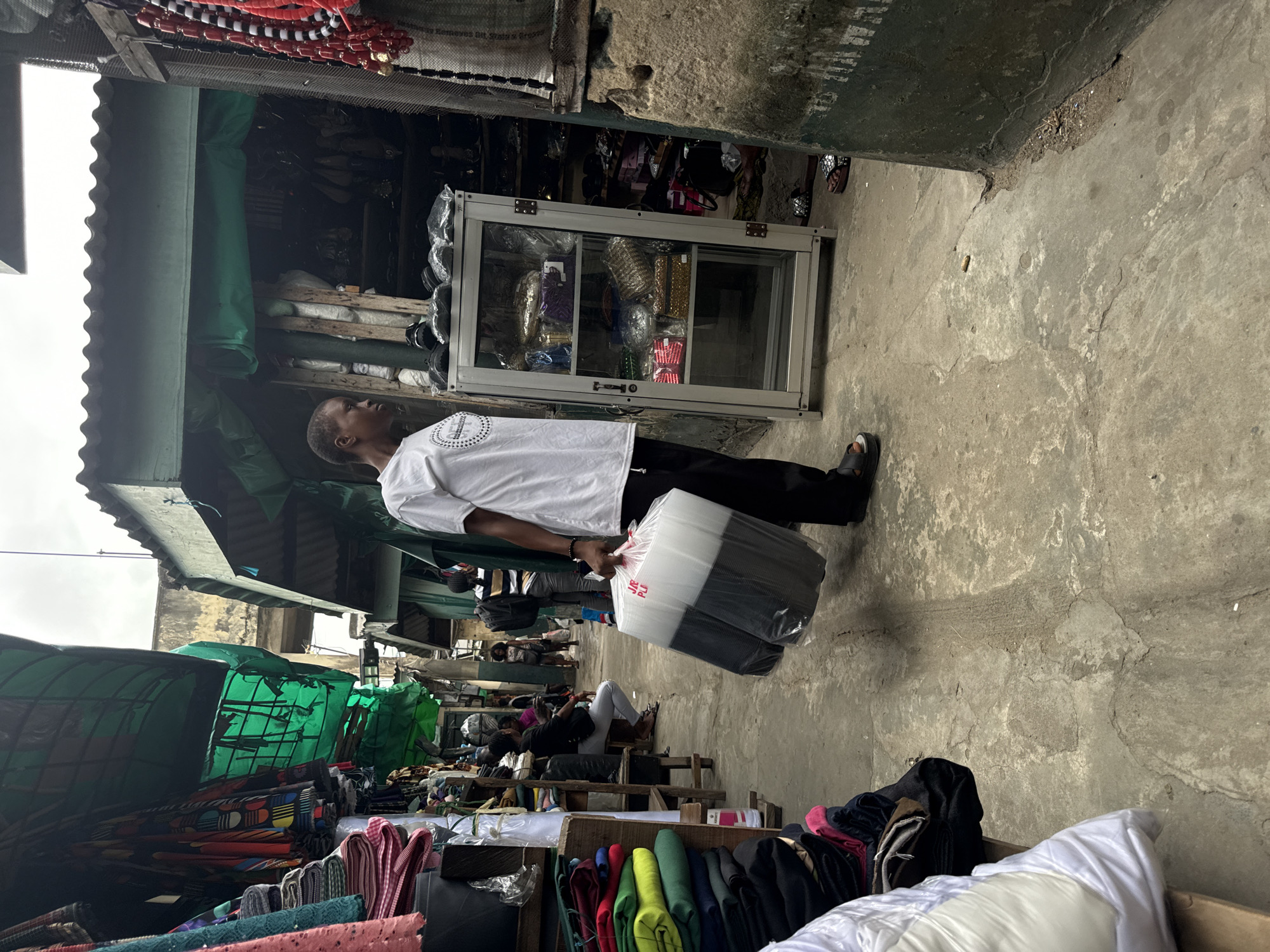 The image depicts a bustling market scene in Lagos, Nigeria. A person, likely a vendor or shopper, is walking through a narrow aisle, carrying a large, clear plastic bag containing black and white items. The aisle is lined with stalls selling various goods, including textiles, clothing, and what appear to be handbags and accessories displayed in a glass cabinet. The environment suggests an outdoor or semi-enclosed market setting, with corrugated metal roofing and weathered structures. Other people are visible in the background, further emphasizing the lively atmosphere of the market. The ground is paved with concrete, showing signs of wear and tear. The overall impression is one of a vibrant commercial hub in the city.