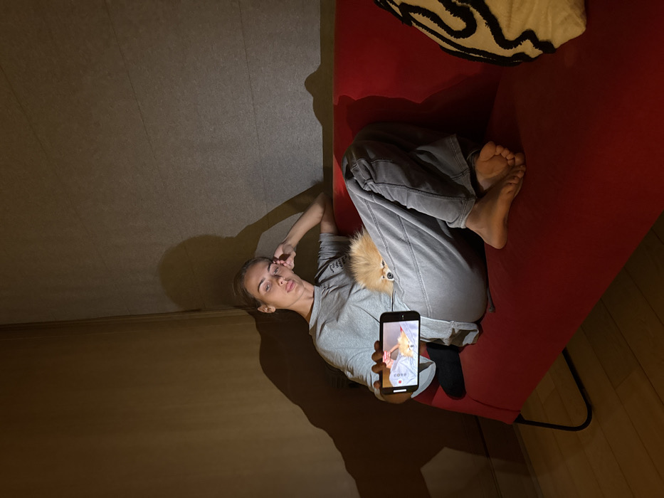 A young adult female is seated on a red upholstered sofa in a dimly lit residential room in Warsaw, Poland. She wears a grey t-shirt and grey sweatpants; her left foot is covered by a black sock, and her right foot is bare. She leans into the corner of the sofa, with her left arm resting on the backrest, and looks directly at the camera with a neutral expression. In her right hand, she holds a black smartphone horizontally, its screen illuminated. The screen displays a video featuring a person's hands interacting with a small, light-colored animal against a blue background, with a "REC" indicator and "PROD" text visible on the display. A small, fluffy, light brown dog, appearing to be a Pomeranian, is nestled between the woman's torso and her bent right leg, with its head visible and facing forward. Distinct shadows are cast on the light brown walls behind the woman due to the phone's light source. A white decorative cushion with black abstract patterns rests on the right side of the sofa. A light-colored wooden floor is visible in the lower left foreground.