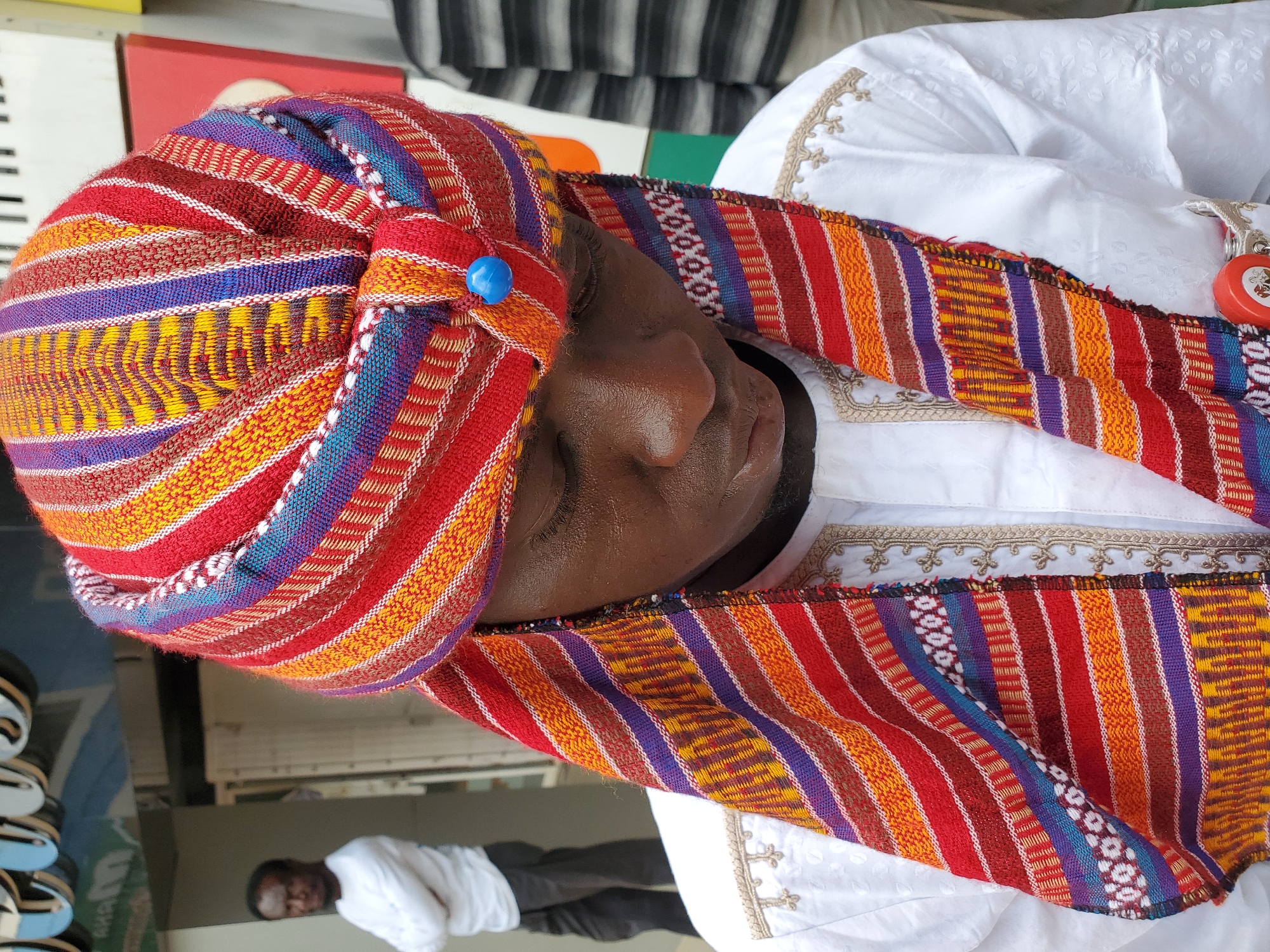 A close-up, portrait-oriented image captured in Jos, Nigeria, features a person, likely male, adorned in traditional West African attire. The individual's head is wrapped in a vibrant, intricately woven fabric that serves as a headwrap or turban. This headwear displays a rich palette of horizontal stripes, including dominant shades of red, orange, yellow, blue, purple, and white, with complex patterns embedded within the bands. A distinctive bright blue spherical bead or button is prominently affixed to the front of the headwrap.
The person's face is seen in profile, with their eyes gently closed, suggesting a moment of repose, contemplation, or perhaps prayer. Their skin tone is dark. They are dressed in a pristine white garment, possibly a kaftan or similar traditional tunic, which is embellished with elaborate gold or beige embroidery around the collar or neckline. A small, circular red object, possibly a keyring or decorative item, hangs near their right shoulder.
In the blurred background, another person, also male, is visible, wearing a white shirt and dark trousers, standing at a distance. Other background elements are indistinct but include what appears to be stacked footwear or sandals on the left and striped fabric (black and white) at the top. The overall scene conveys a sense of cultural heritage and quiet dignity, possibly during a ceremonial event or social gathering. There is no visible text in the image.