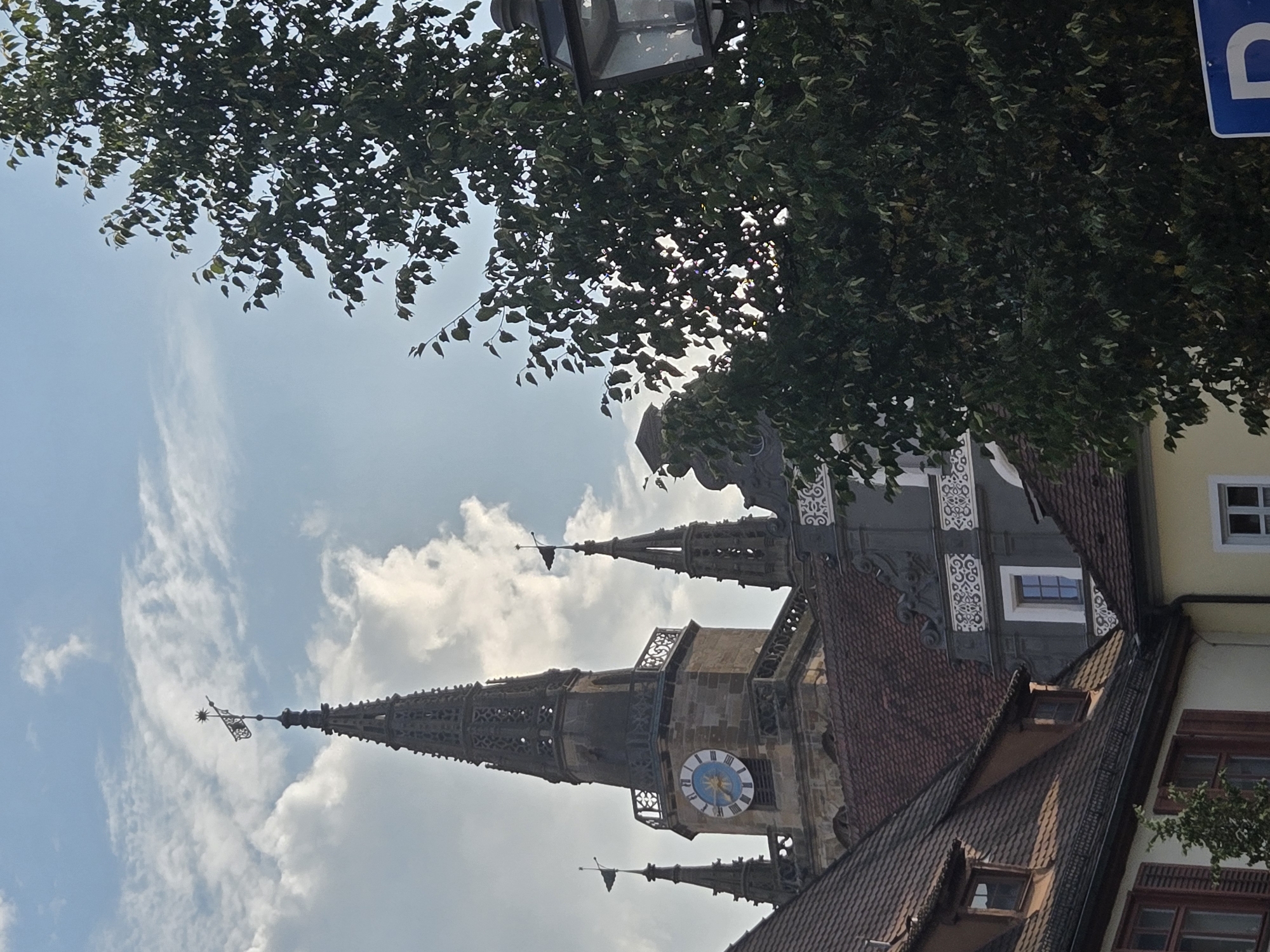 The image captures a view of the Ansbach Town Hall (Rathaus), a prominent historical building in the city of Ansbach, Germany. The focus is on the tower and parts of the surrounding architecture. The tower, constructed of stone, features intricate stonework and ornate detailing, particularly on its spire. A large clock face, with a blue and white patterned dial, is visible on the tower. Two smaller spires, adorned with flags, flank the main tower.
In the foreground, to the right, is a large, leafy tree, its branches obscuring parts of the sky and the building. The leaves are a rich green, suggesting it's likely a temperate season. Peeking out from behind the tree on the far right is a street lamp with a glass lantern.
The background consists of a bright, partly cloudy blue sky. The lower section of the image reveals the red-tiled roofs of adjacent buildings, with visible windows and architectural embellishments on one of the structures. The scene depicts a picturesque urban landscape, likely taken from a street level perspective, looking upwards towards the historic town hall. No people or specific actions are depicted.