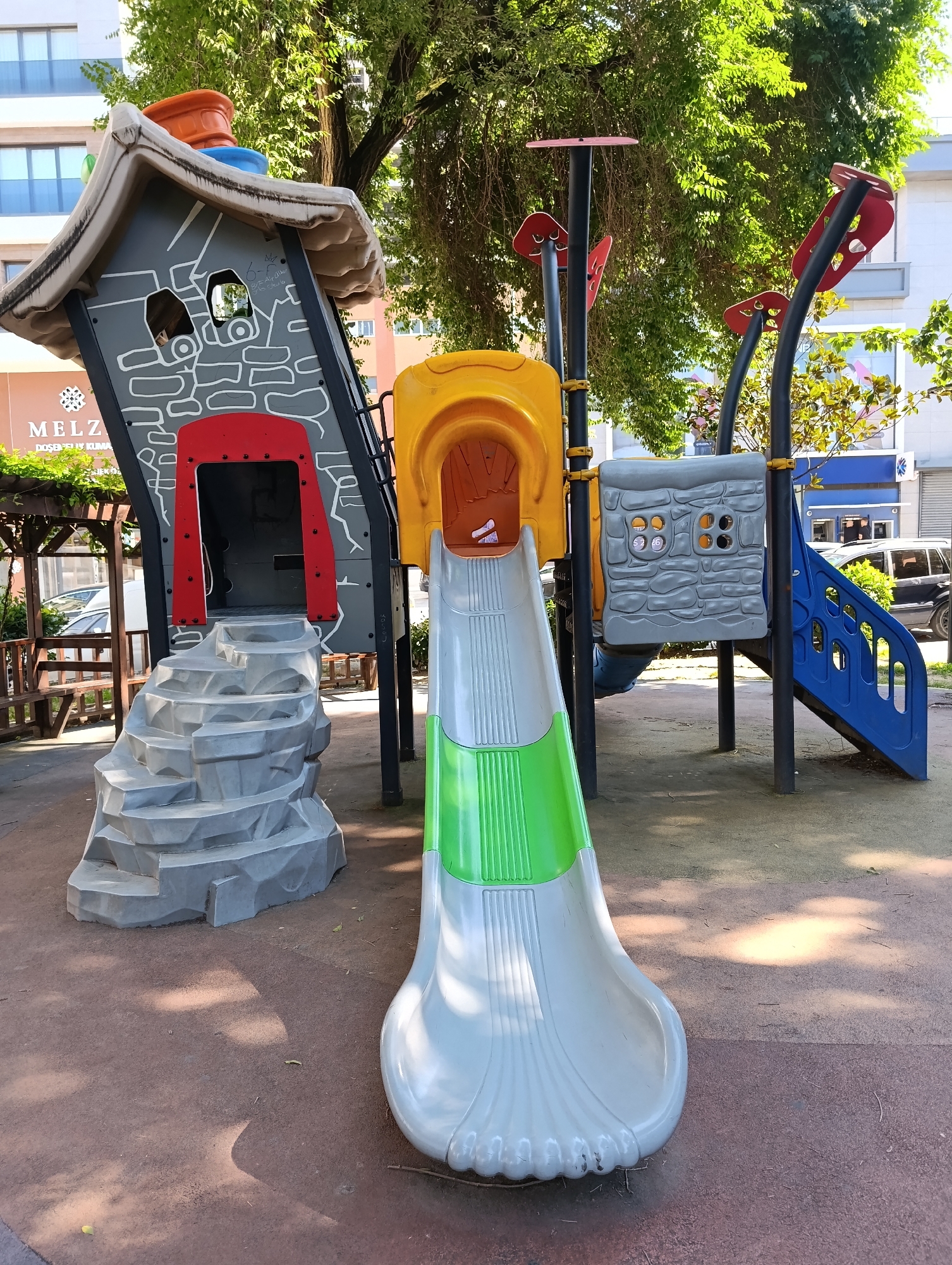 The image shows a children's playground in Bayrampaşa, Turkey, with a multi-colored slide, a climbing structure resembling a house with a red door and gray stone pattern, and a blue slide on the right. The playground surface is a mixture of red rubber and brown soil, with scattered leaves. In the background, there are trees, buildings, and a parked black SUV. The lighting suggests it is daytime with sunlight filtering through the trees, creating dappled shadows on the ground. There are no people visible in the image.