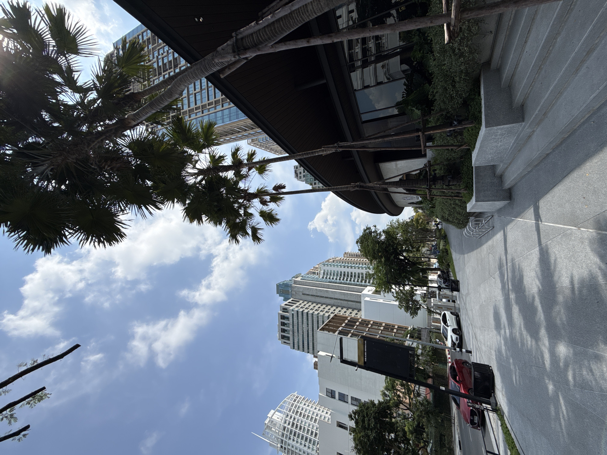 The image shows a street scene in Bangkok, Thailand. Tall buildings of different architectural styles are visible. The sky is blue with white clouds. Several palm trees line the street. A modern building with a dark brown roof is also present. Parked cars are on the right side of the street. A grey sidewalk with stairs is on the right. Trees and greenery are on both sides of the street.
