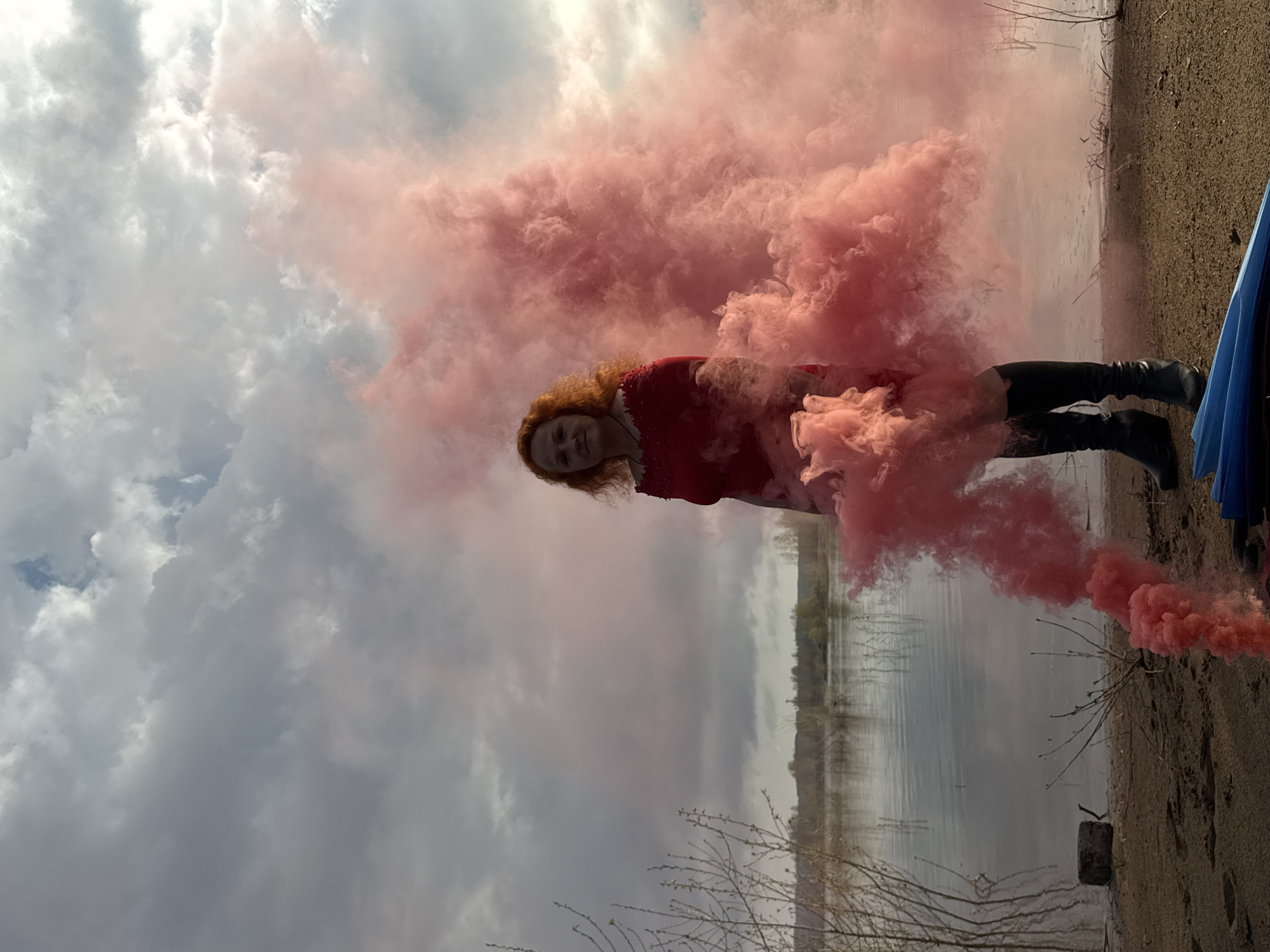 A woman with reddish-brown hair is the subject of this outdoor photograph, surrounded by a cloud of pink smoke. She is wearing a red dress and black boots, standing on what appears to be a sandy shore.
In the background, there is a body of water, likely a lake or river, with the opposite shore visible. The sky is overcast with grey and white clouds, suggesting an overcast day.
The location is likely near a body of water in Noviy, Russia, given the description of the environment. The photo is taken at an angle, with the ground appearing at the top and the sky at the bottom, altering the traditional perspective.