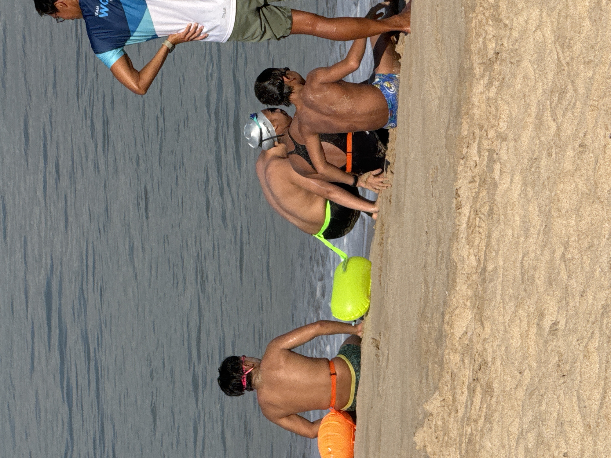 The image depicts a group of individuals on a sandy beach in Kuta, Indonesia, near the water's edge. The ocean's surface is calm with gentle waves. In the foreground, three young people are seated on the sand. The person on the far left is a male, facing away from the camera, wearing a pink headband and green swim trunks. To his right, another individual, likely also male and wearing a black swim cap and goggles, is seated, facing slightly away. Between them is a bright green inflatable buoy. Further to the right, a third person, a young male, is seated and facing away, wearing blue patterned swim trunks. A fourth person, a male, stands to the right of the seated individuals, facing left, wearing a blue and white sports shirt and green shorts. Two orange inflatable buoys are positioned to the left of the seated individuals. The scene suggests a training or recreational activity related to swimming or water sports, given the presence of the buoys and swim gear. The lighting indicates it is daytime.