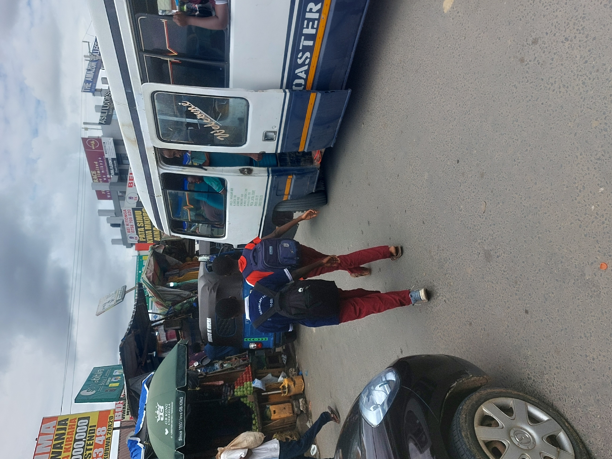 Here is a detailed description of the image:
The image shows a street scene in Dar es-Salaam, Tanzania. Two young people in school uniforms are walking on the side of the road. A white bus with blue stripes labeled "COASTER" has its door open, with people inside. Several market stalls and shops line the street, selling various goods, and there are signs for businesses like "Somabiblia Book Center" and advertising "GB KING Umbrella". A black car is parked on the right side of the image, partly blocking the view. The sky is cloudy. The bus is in front of several other stores and advertisements. The location appears to be a busy commercial area with a lot of pedestrian and vehicular traffic.