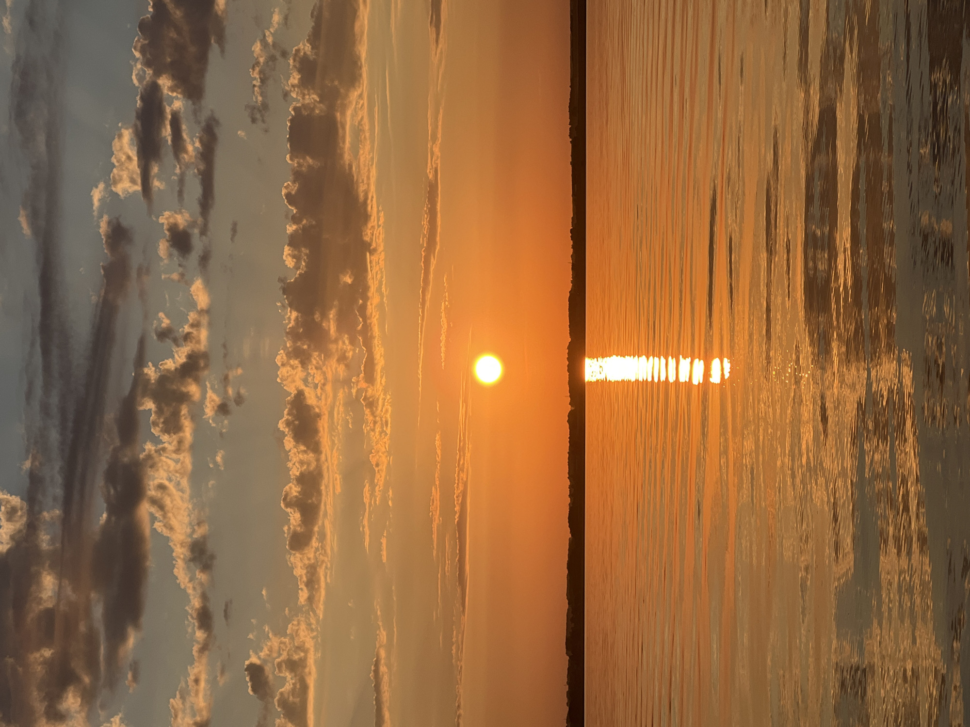 The image depicts a serene natural landscape, likely a body of water such as a lake or a wide river, bordering a treeline under a sunset sky. The sun, a bright orb, is positioned slightly to the left of the frame, casting a brilliant golden-orange glow across the scene. Its reflection shimmers intensely on the rippling water surface, creating a striking vertical path of light. The sky is filled with a mix of soft, wispy clouds tinged with warm hues of orange and yellow, contrasting with darker, more textured clouds higher up. The horizon line is clearly defined by a dark silhouette of trees and possibly some distant structures. The water appears calm, with gentle waves creating horizontal patterns and reflecting the colors of the sky.
The location is identified as Novopichugovo, Russia. However, there are no visible human elements, buildings, or specific urban features that would distinctly place this scene within a city. It is purely a natural, scenic view during sunset.