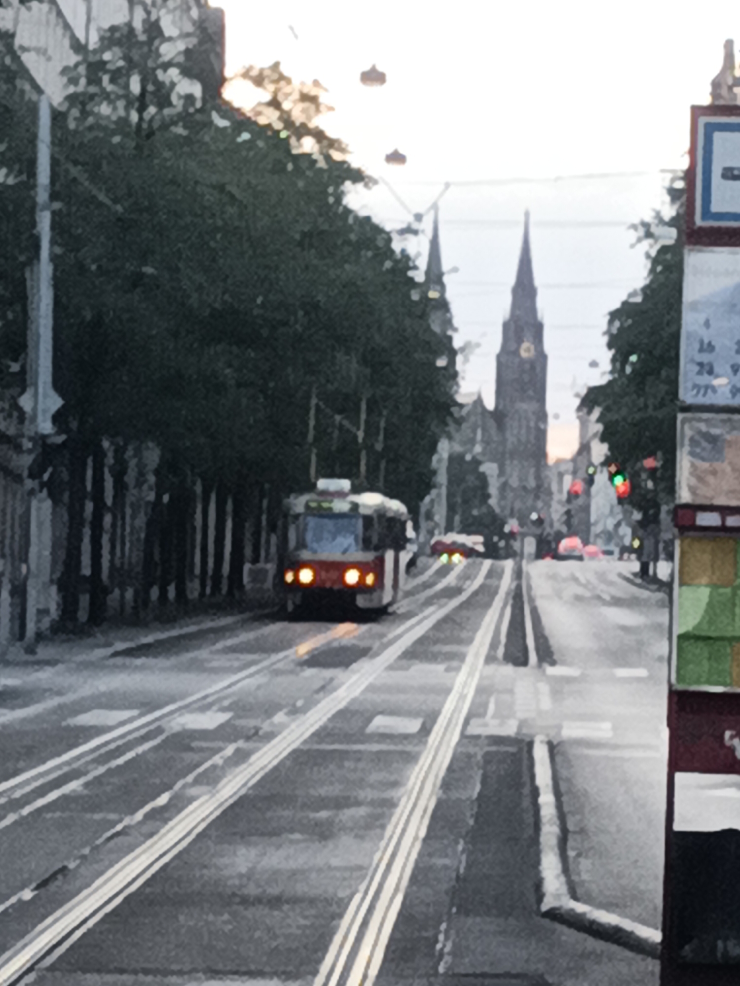 An outdoor, daytime, eye-level, full shot shows a street in Prague, Czechia. The street is lined with trees on the left and buildings with spires in the background. A tram is visible on the street. The tram is red and white and has its headlights on. The street is wet and reflects the lights of the tram and streetlights. There are tram tracks on the street. A signpost with a timetable is on the right side of the image. The sky is overcast.