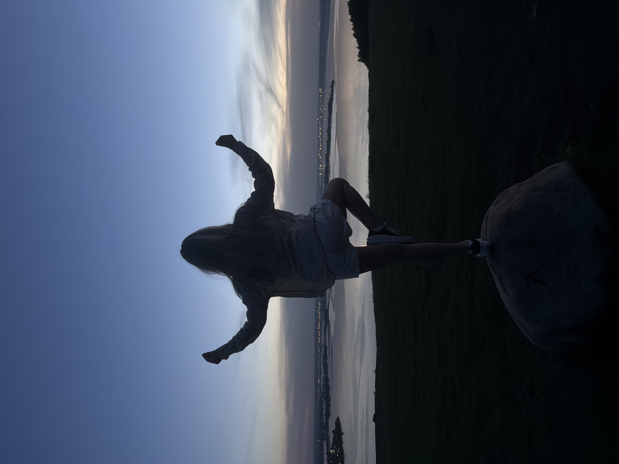 A person is sitting on a rock with their legs crossed, in front of a view of the city of Innopolis, Russia at twilight. The person is wearing a light-colored top, shorts and shoes. Their arms are raised. The background consists of a body of water and the city lights of Innopolis reflected on the water. The sky is a gradient of blue and grey. There is some land on the right side of the image, and the person is sitting on a rock in front of this area.