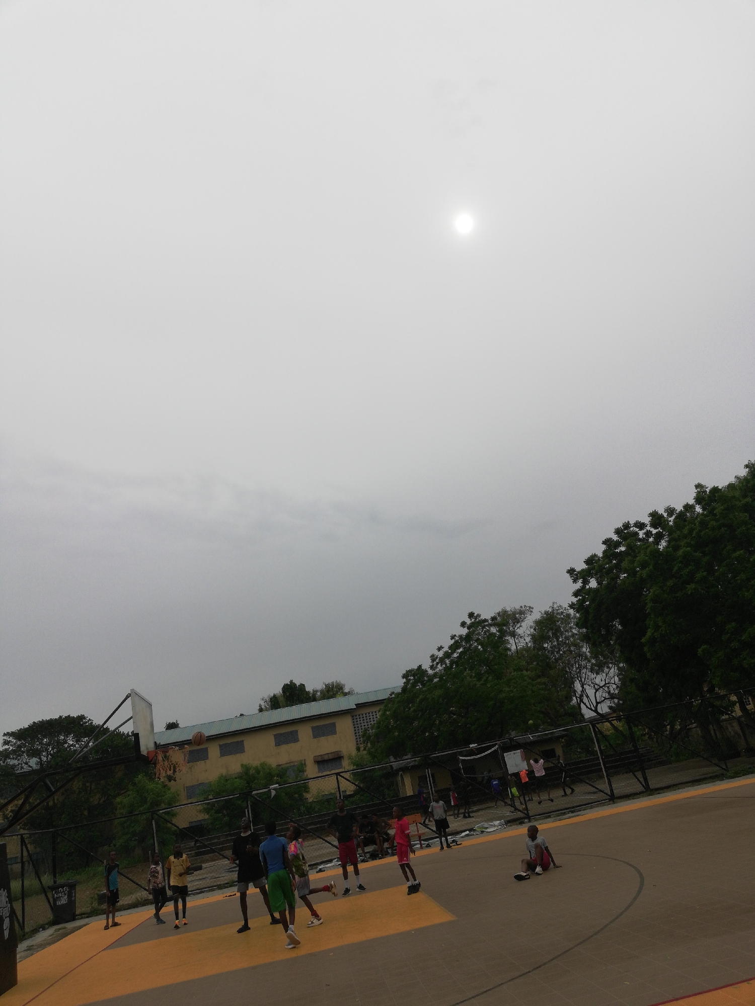 The image depicts an outdoor basketball court in Lagos, Nigeria. Several young people are playing basketball, while another is sitting on the court. The court is fenced, with trees visible beyond the fence. A building with windows is visible in the background. The sky is overcast. A basketball is in the air near one of the hoops.