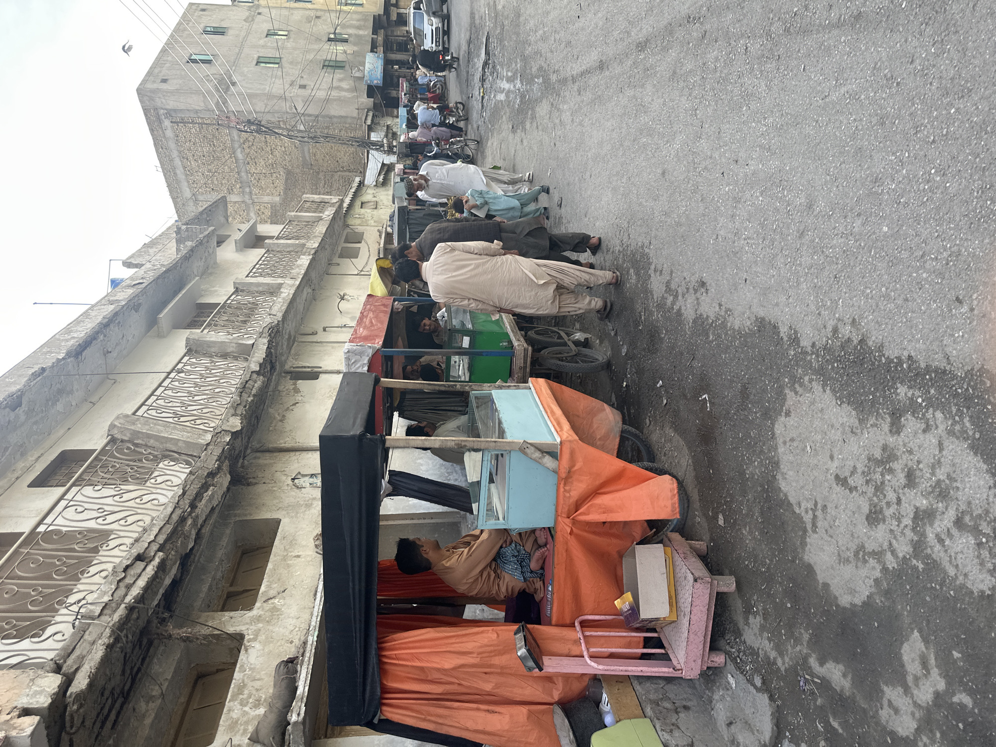 The image depicts a vibrant street scene, likely in a South Asian urban area. On the left, several multi-story buildings line a narrow, paved road. These structures appear aged, characterized by exposed concrete, some crumbling facades, and ornate yet worn metal railings on balconies.
Along the roadside, a group of men, mostly dressed in traditional shalwar kameez, are gathered. Some are standing, seemingly conversing, while others appear to be waiting or observing. A small child is also visible among the adults.
In the foreground and midground on the left, two makeshift carts stand out. One cart, covered with orange fabric and featuring pink metal framing, serves as a temporary shelter or stall. Inside this cart, a man in a light brown outfit is seen lying down, resting. Behind it, another cart with a green base is visible, and another man is sitting inside it.
Further down the street, additional people, parked vehicles including a white van, and bicycles can be discerned. The sky above is bright and overcast, and numerous utility lines crisscross the view overhead. On a small cardboard box placed on the pink cart, the word "LUCKY" and parts of another word, possibly "MINT," are visible. The scene portrays a typical daytime routine in a bustling, well-inhabited street.