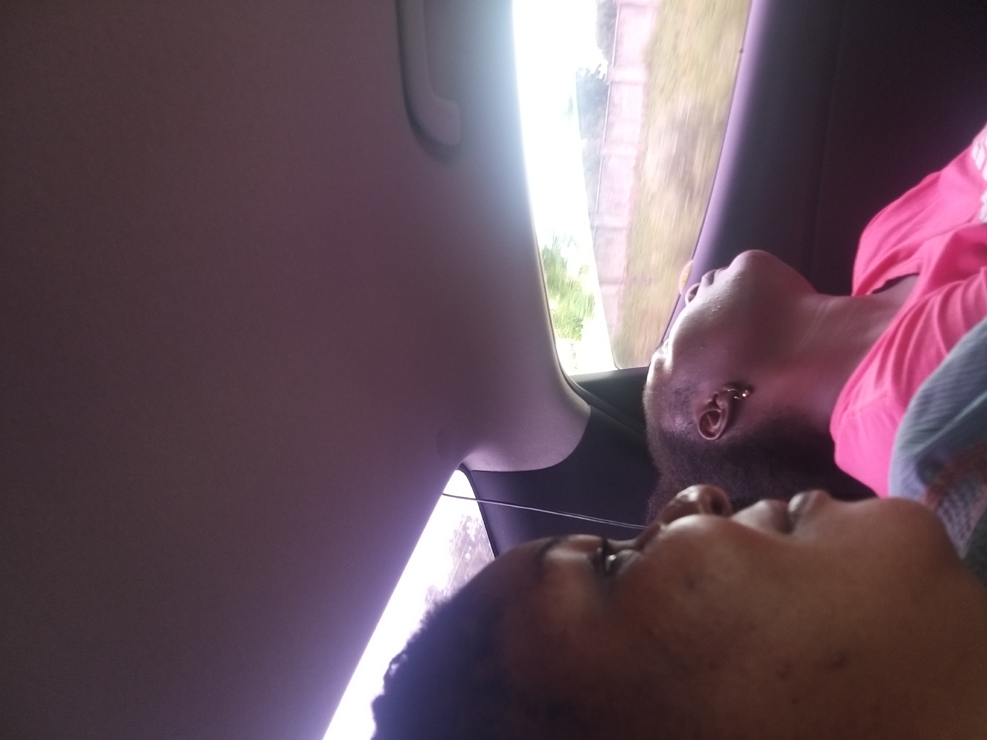 This is a close-up, eye-level, indoor shot taken from the back seat of a vehicle, likely in Kafarati, Nigeria. The image features two individuals. On the left, a person's face is partially visible, turned to the right, with their gaze directed off-frame. Their skin appears dark, and they are wearing a dark garment. To the right, another individual is seen from the shoulders up, facing away from the camera and towards the right side of the frame. This person appears to be asleep, with their head tilted back and eyes closed. They have dark hair and are wearing a bright pink t-shirt and a small hoop earring in their visible ear. The background shows the interior of the car, including the headliner and a car door. Outside the car window, a blurred landscape with a concrete wall and vegetation is visible, suggesting movement. The lighting is bright, particularly from the window, casting a purplish hue on the interior surfaces.