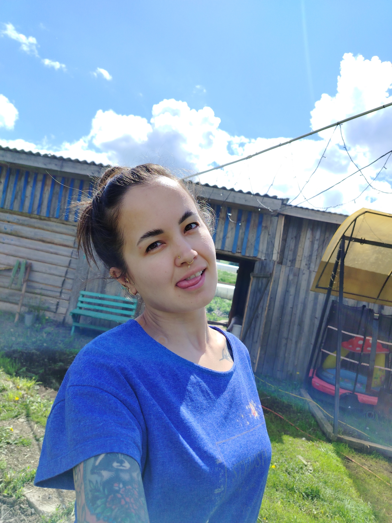 The image is a selfie of a woman with fair skin and dark hair pulled into a messy bun, standing outdoors in Гожан, Russia. She is wearing a blue t-shirt and has a visible nose ring and earrings. She is sticking her tongue out. A colorful tattoo is visible on her left arm.
Behind her is a weathered wooden structure with a blue-striped section. There is a green bench and a translucent covered structure to the right of the woman. Scattered green vegetation and dandelions populate the ground. Overhead, a partly cloudy blue sky is visible. Strands of wire stretch across the scene. A faint haze or smoke is visible in the lower-left corner.