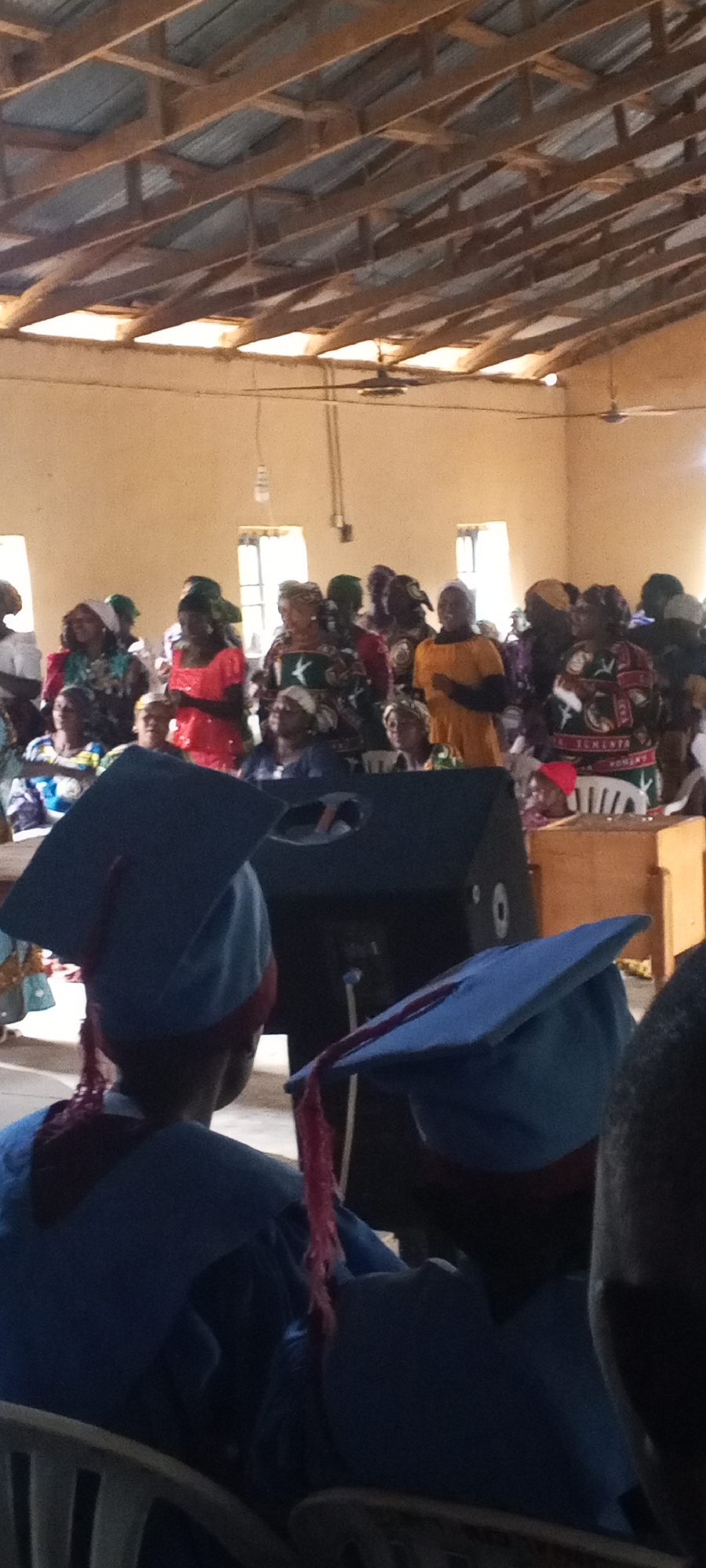 The image depicts an indoor gathering in Bahit, Nigeria, possibly a ceremony or a service. The environment appears to be a hall with a wooden truss ceiling and corrugated metal roofing. Two ceiling fans are visible. In the foreground, two individuals are wearing graduation caps and gowns, with the caps being light blue and gowns a darker blue. The individuals in the background are dressed in colorful traditional attire, predominantly women, some wearing headscarves. One woman in the background is wearing an orange outfit. A large black speaker is positioned centrally in the mid-ground, with a wooden box-like object to its right. The lighting is natural, coming from windows on the back wall. The overall scene suggests a community event, possibly a graduation ceremony for a local school or organization.