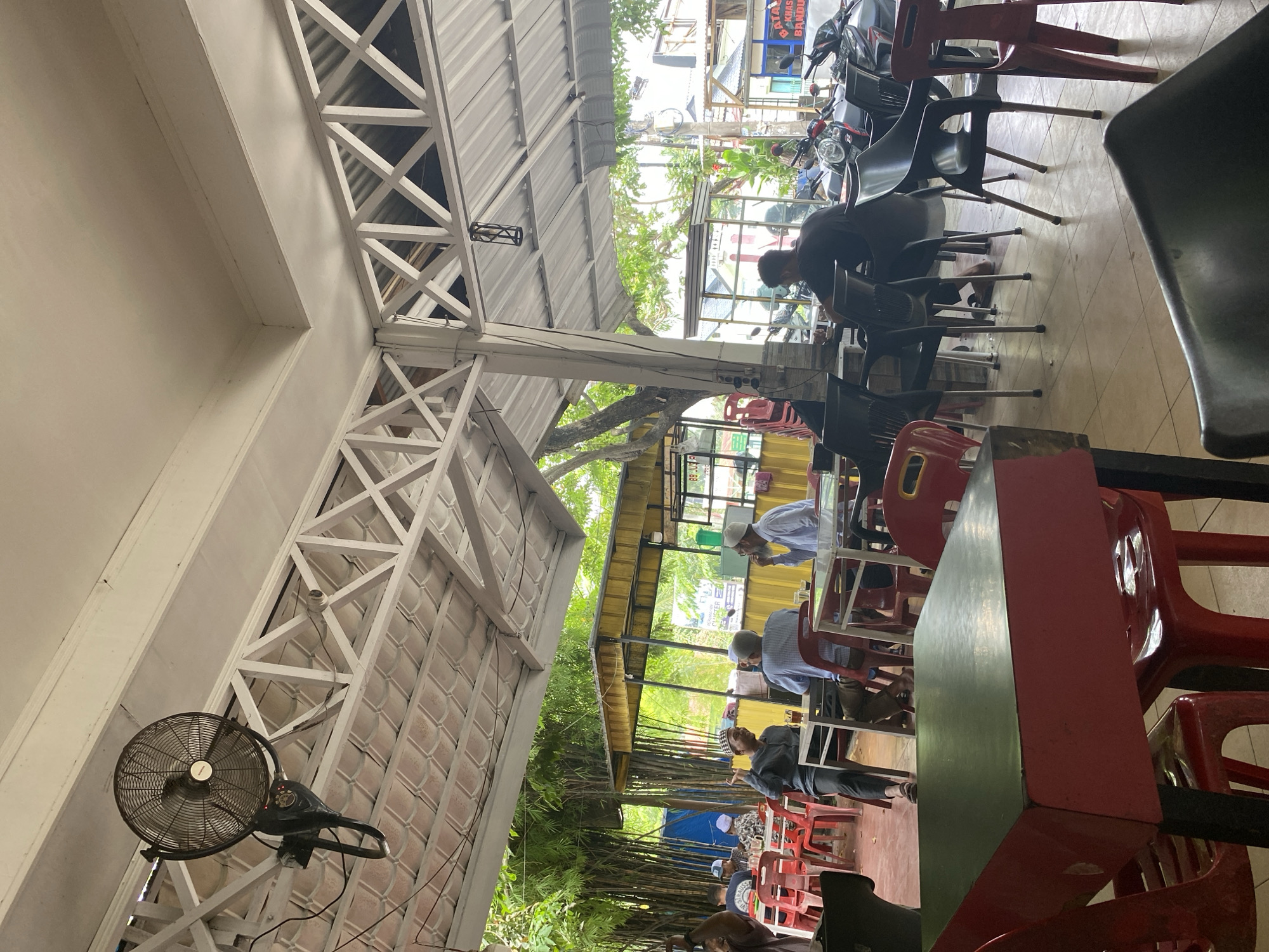 The image depicts an outdoor restaurant in Lhokseumawe, Indonesia. The restaurant is covered with a corrugated metal roof supported by white-painted steel trusses. A black fan is mounted to one of the trusses. Patrons are seated at red and black plastic chairs and tables. Several individuals are visible, some wearing Muslim attire, suggesting a possible religious context. A man in a black shirt works on a motorcycle parked near the restaurant. Beyond the restaurant, there is greenery and bamboo trees. The setting is a typical Indonesian open-air eatery.