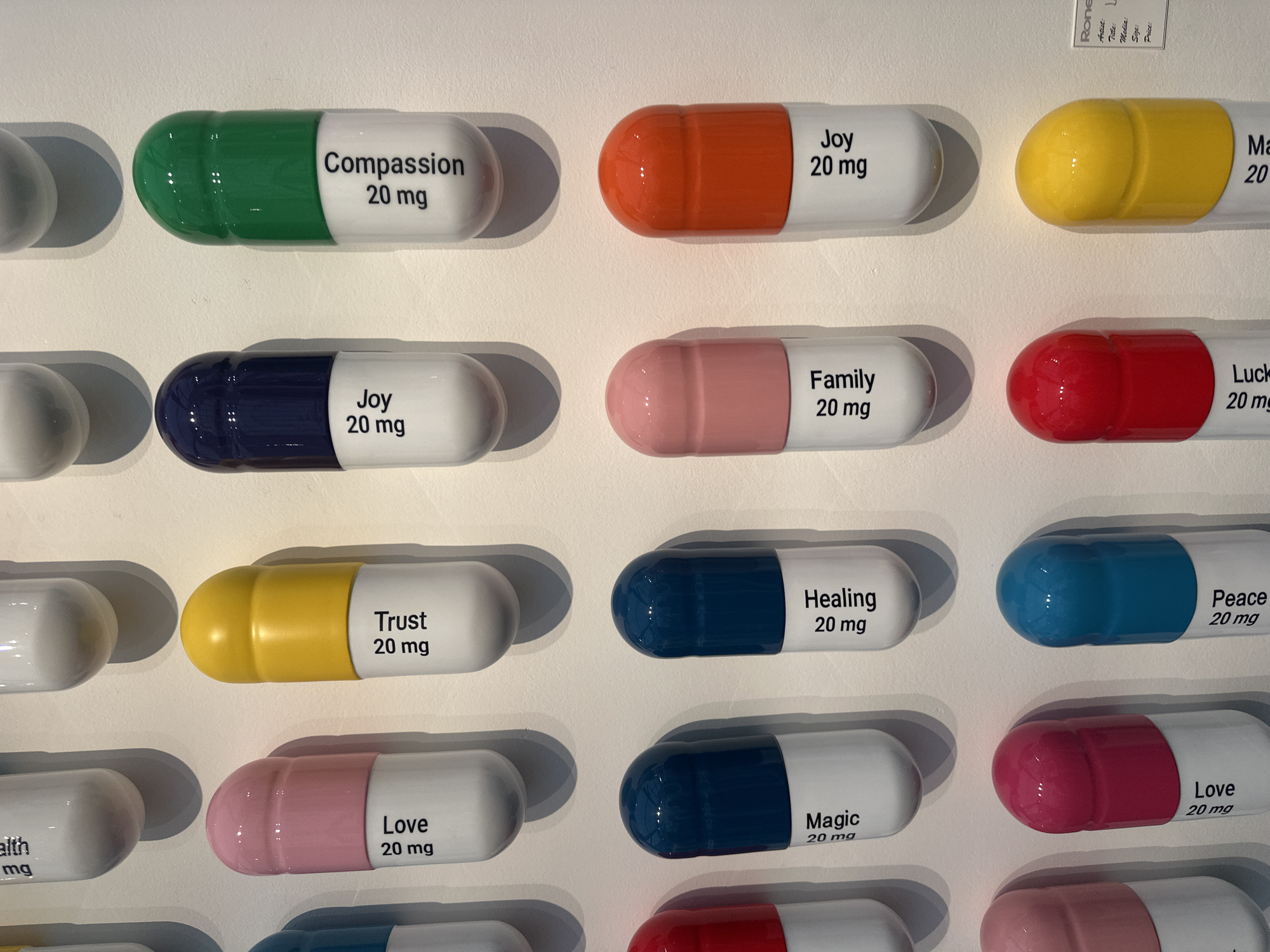 The image shows a close-up of a piece of artwork featuring rows of colorful, oversized pill-shaped objects attached to a white wall. Each "pill" has a different color on one end and white on the other, with text printed on the white section. The texts are positive words associated with well-being, such as "Compassion 20 mg," "Joy 20 mg," "Family 20 mg," "Trust 20 mg," "Healing 20 mg," "Love 20 mg," and others. The artwork is well-lit, and the pills cast subtle shadows on the wall. A fragment of a label with text "Rone", "Title", "Media", "Size", and "Price" can be seen on the top-right corner. No people are visible in the image.