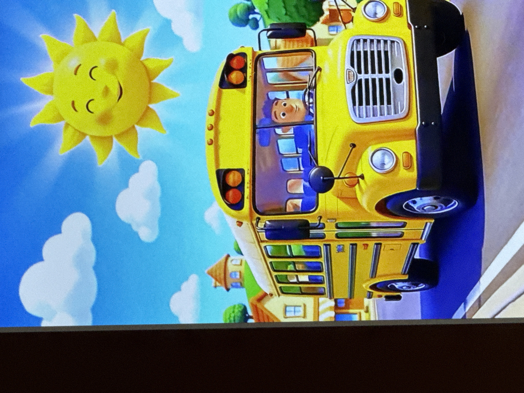 A cartoon illustration depicts a bright yellow school bus driving down a street. A smiling cartoon sun with rays shines in a blue sky with white clouds. The bus has a cartoon driver with purple hair behind the wheel. In the background, there are stylized houses with orange roofs and green trees. The scene is vibrant and cheerful. There is no indication of the location being the city of Cahul, Moldova, as this appears to be a fictional, animated setting.