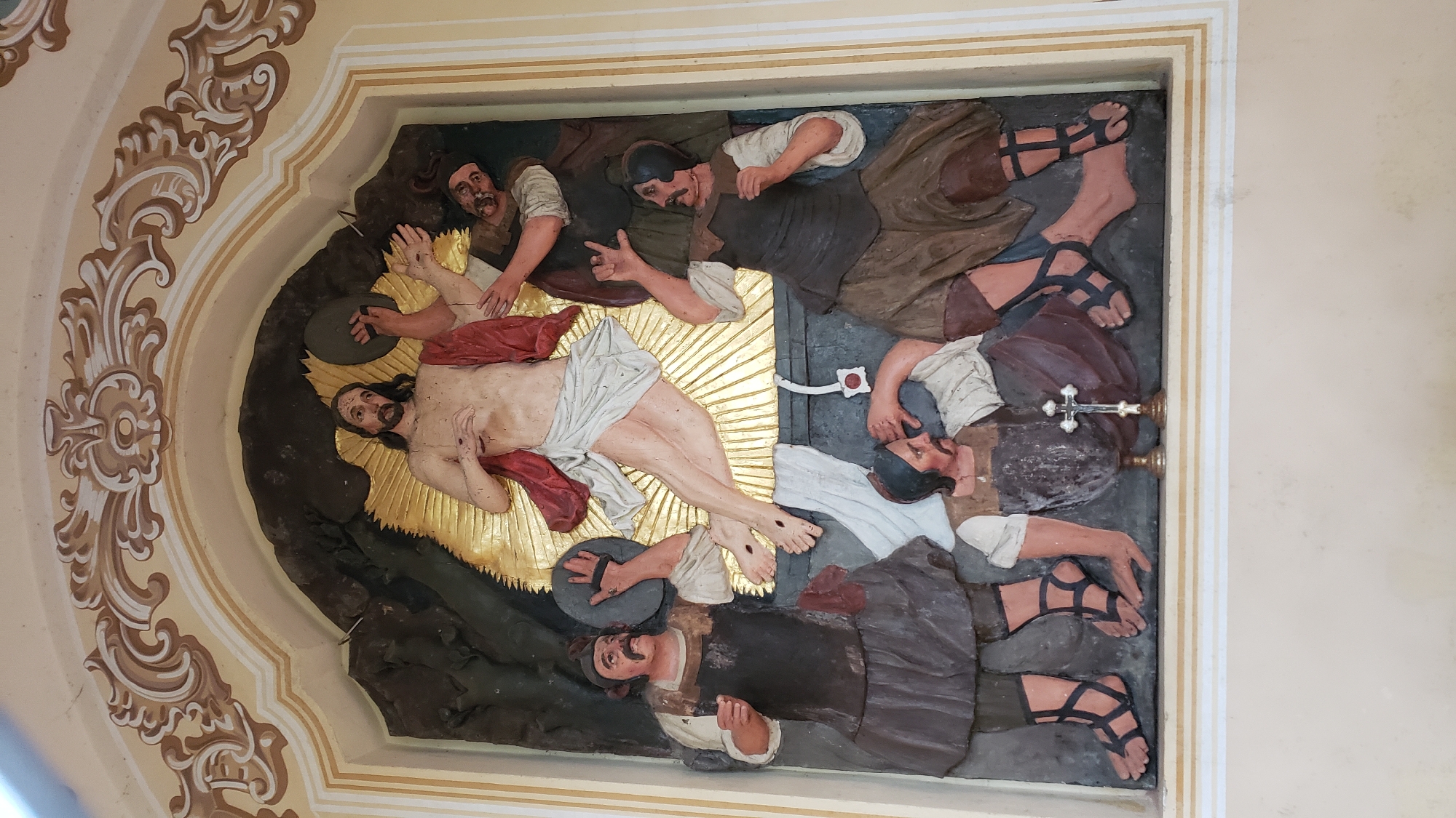 This is a photograph of a carved relief depicting a religious scene, likely the Crucifixion or a related event. The central figure is Jesus Christ, lying on his back with his body stretched out. He is depicted with a beard, long hair, and a loincloth. His skin is pale, and his wounds are visible, including marks on his hands and feet.
Surrounding Jesus are several other figures, presumably Roman soldiers or mourners. Some of these figures are shown in various states of action or repose. One man to the left of Jesus is holding a circular object, possibly a hammer or a plate. Another man is positioned above Jesus, his hand reaching towards him. Further to the right, several figures are lying down or seated, some appearing to be in distress or deep in thought. One figure in the foreground has a mustache and is wearing a tunic and trousers.
The figures are rendered in a stylized, somewhat primitive manner, with expressive faces and poses. They are clothed in tunics, trousers, and sandals. The scene is set against a dark, textured background, with a bright, golden halo or sunburst emanating behind Jesus. The relief is set within an architectural niche, framed by ornate, decorative moldings in beige and brown tones, characteristic of Baroque or Rococo style.
The overall impression is one of drama and solemnity. The expressions on the faces of the figures suggest a range of emotions, from pain and sorrow to perhaps a sense of duty or indifference. The lighting in the photograph highlights the three-dimensionality of the carvings. The location context of Prešov, Slovakia, suggests this relief is likely part of a church or religious building. No visible text is present in the image.