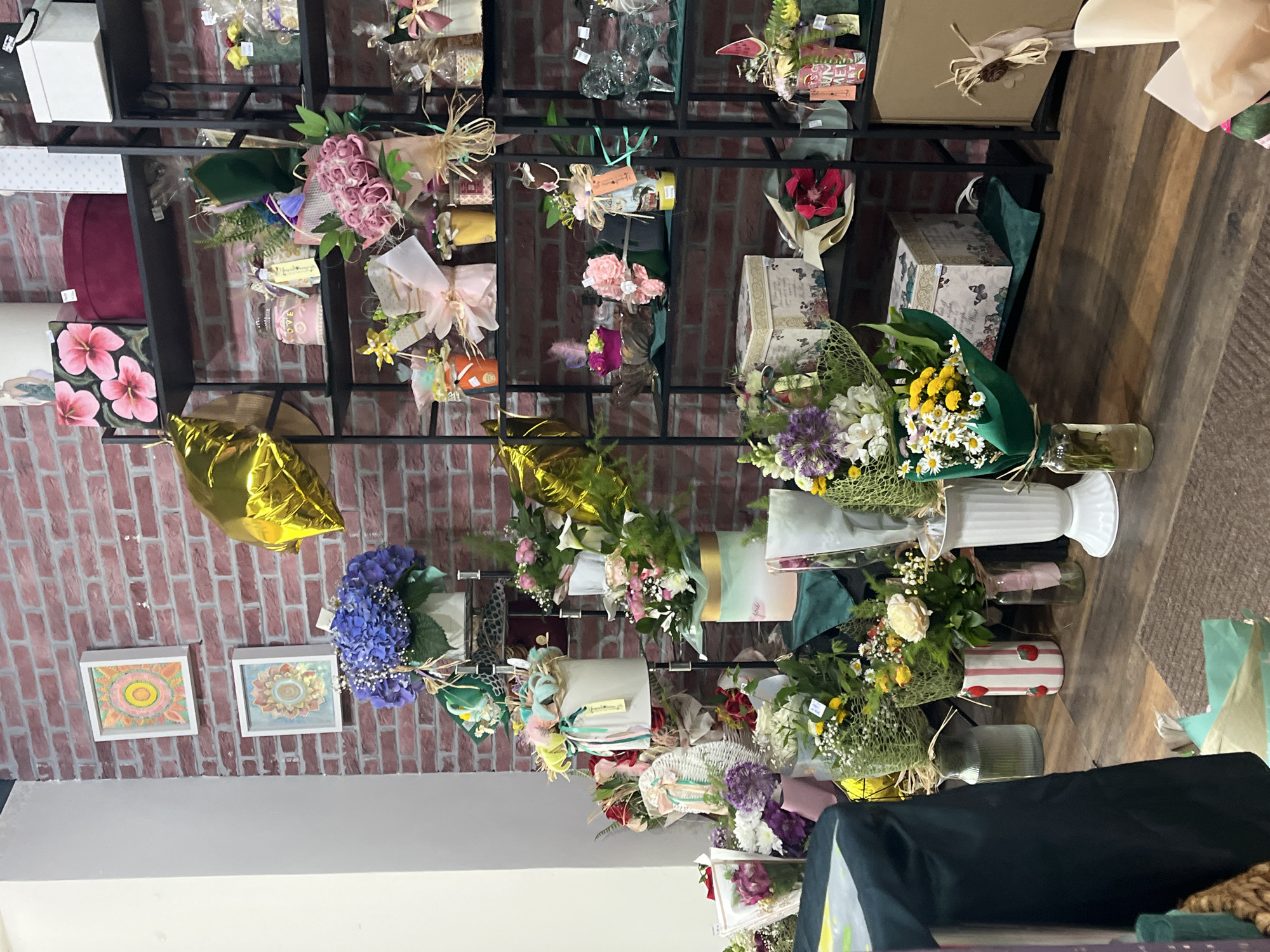 The image depicts the interior of a flower shop or a shop that sells floral arrangements. The location is inferred to be in Burgas, Bulgaria, although this is based on external knowledge and not explicitly shown in the image.
The shop has a brick wall backdrop where various floral arrangements and gift items are displayed on metal shelving units. The arrangements include bouquets, boxed flower sets, and decorative items, often wrapped in cellophane or adorned with ribbons and other decorative materials. The flowers have a wide range of colors, with pink, purple, white, and yellow flowers being prominent. Several gold balloons and framed art pieces are also visible.
The floor is made of dark wooden planks. There is a table covered with a dark cloth, possibly used for preparing or displaying items. Vases with flowers are placed on the table.
The general impression is that of a well-stocked retail space specializing in floral gifts, with a varied inventory and a decorative presentation.