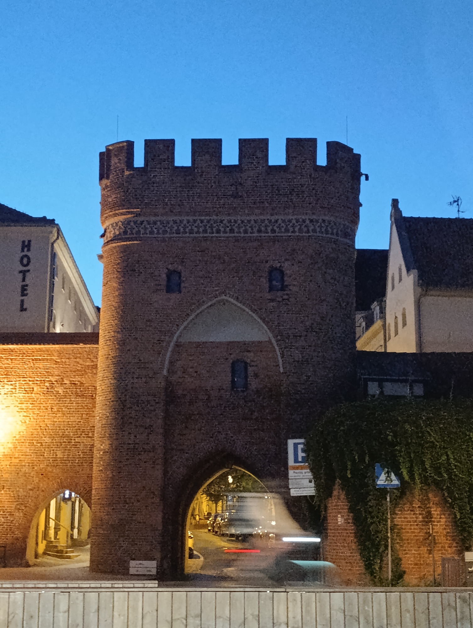 The image shows a brick gate tower in the city of Toruń, Poland. The tower has a crenellated top and an arched entrance. A wall extends from the tower, also made of brick and illuminated by warm lights. To the left of the tower, a building with the word "HOTEL" in large vertical letters is visible. To the right of the tower, lush green ivy covers a brick wall. Through the arched entrance of the tower, a street with cars can be seen, with some light trails indicating movement. The sky is a deep blue, suggesting it is either dusk or dawn. A street sign with a blue "P" for parking and a triangular sign with a silhouette of a person are also present near the ivy-covered wall. The scene depicts a historic architectural element within a modern urban setting.