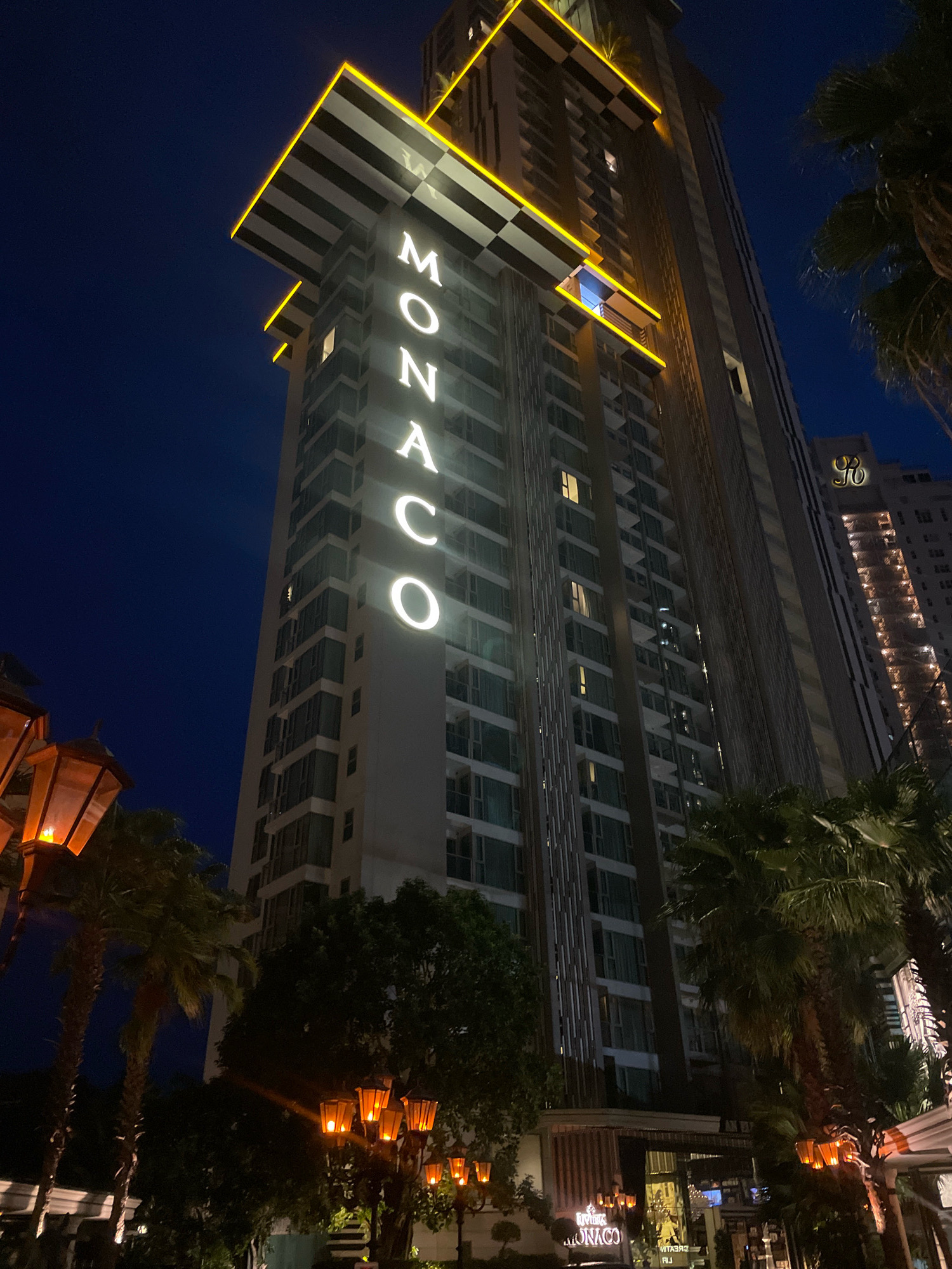 The image shows a tall, modern building at night, likely a hotel or condominium, with the word "MONACO" prominently displayed in large white illuminated letters on its facade. The building features a distinctive architectural element at its top with black and white striped banding and yellow neon lighting. The scene is set in Pattaya City, Thailand, as indicated by the presence of palm trees and the overall ambiance of a tropical city at night. Several outdoor lamps with orange light cast a warm glow on the tropical foliage. In the background, another tall building with illuminated windows is visible, along with a sign that appears to be "R" in a decorative font. The sky is a deep, dark blue, typical of nighttime. There are no people clearly visible in the image.