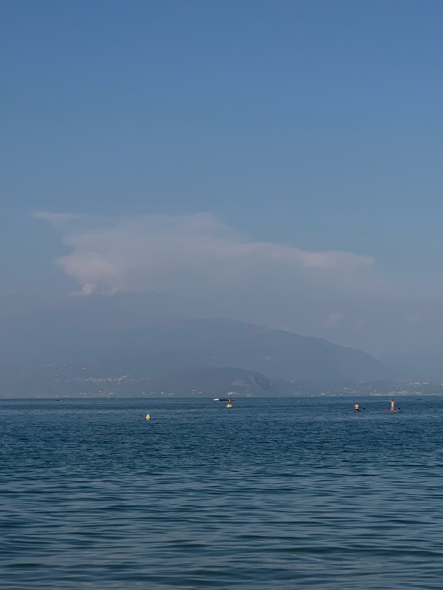 A wide shot captures the tranquil waters of Lake Garda, viewed from Padenghe sul Garda, Italy. The azure sky, a pale blue, is partially obscured by a wispy white cloud formation. In the distance, the soft contours of mountains rise, their slopes dotted with a faint outline of a town or villages. The lake's surface is a deep, calming blue, rippled by gentle waves. A bright yellow buoy floats on the water to the left of the frame. Further out, three individuals are engaged in paddleboarding. One person stands on a board, paddle in hand, while another, closer to the foreground, also stands on a board, seemingly about to paddle. A third paddleboarder is further away and to the right. The scene conveys a sense of peace and leisure.