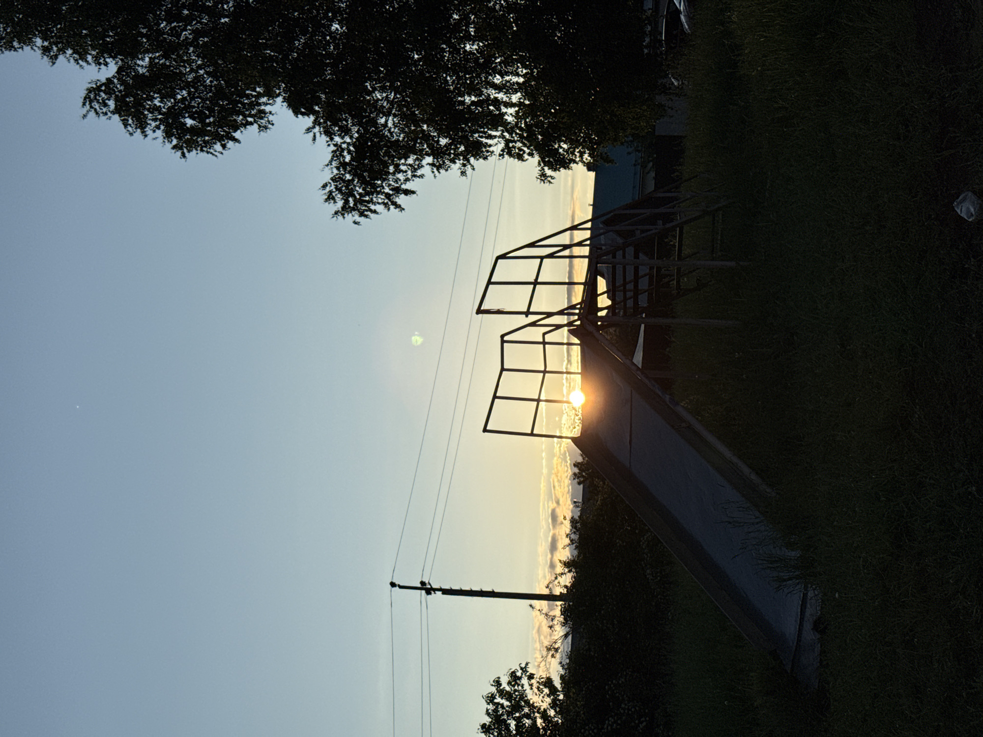 This outdoor photograph, set in Прикамский, Russia, captures a tranquil scene at either sunrise or sunset. The sky dominates the upper portion of the frame, transitioning from a soft blue at the top to vibrant yellows and oranges near the horizon, where the sun is prominently featured. The low sun casts a warm, bright glow, creating high-contrast silhouettes of the foreground elements.
A children's slide, dark and starkly silhouetted, occupies the right side of the image. Its metal railings and stairs lead up to the top, with the sun shining intensely through the structure, causing a bright glare. To the left of the slide, a utility pole with several power lines extends vertically across the midground, also appearing as a dark silhouette against the bright sky. Dark, dense tree foliage frames the top of the image, while dark green grass and bushes cover the lower portion of the frame, surrounding the base of the slide. A small, light-colored object, possibly a plastic bag, is visible on the ground in the far right. No people are present, and no specific activity is taking place. The notable details are the golden hour lighting and the strong silhouetting of objects. There is no visible text.