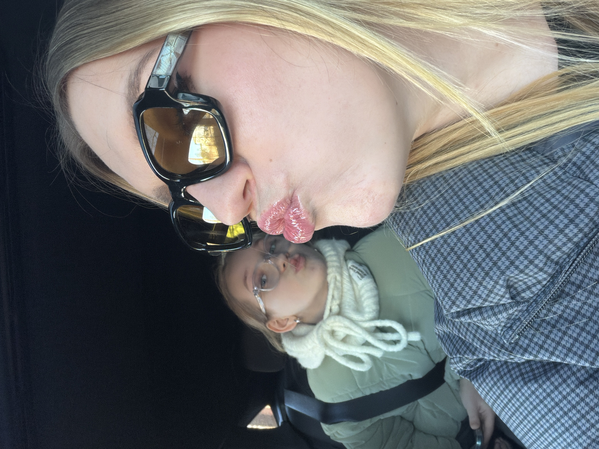 Two blond-haired females are pictured in what appears to be the inside of a vehicle. The face of the older female is in the foreground, tilted at an angle, and she is wearing sunglasses with black frames and tinted lenses. Her lips are pursed as if kissing. She is wearing a gray and black houndstooth patterned garment. The younger female is in the background, also pursing her lips in a kissing gesture. She wears round glasses and has a white scarf around her neck, and a light green puffy jacket. A black seatbelt crosses her chest. The interior of the vehicle is dark. The location within the city of Riga, Latvia is not determinable from the image.