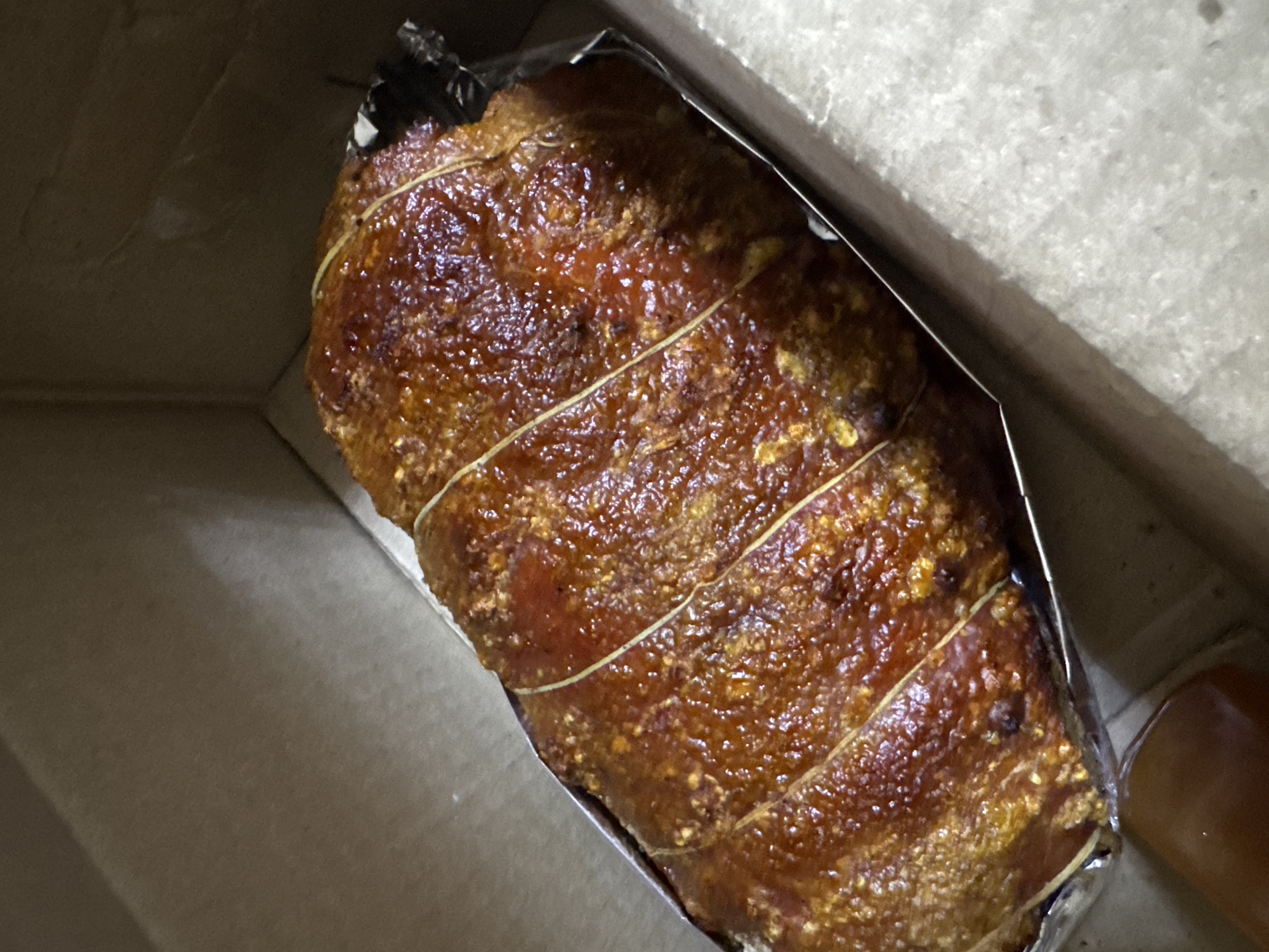 A close-up, slightly angled view depicts a whole roasted pork belly, likely a Lechon Belly, wrapped in foil and tied with string, nestled inside a cardboard box. The pork skin is deep brown and glistening, suggesting it is freshly cooked and crispy. The foil lining conforms tightly to the pork's shape. The inside of the box is a muted beige or gray. The image suggests this might be a take-out order or delivery from a restaurant in Davao City, Philippines, given the popularity of Lechon Belly in the region.