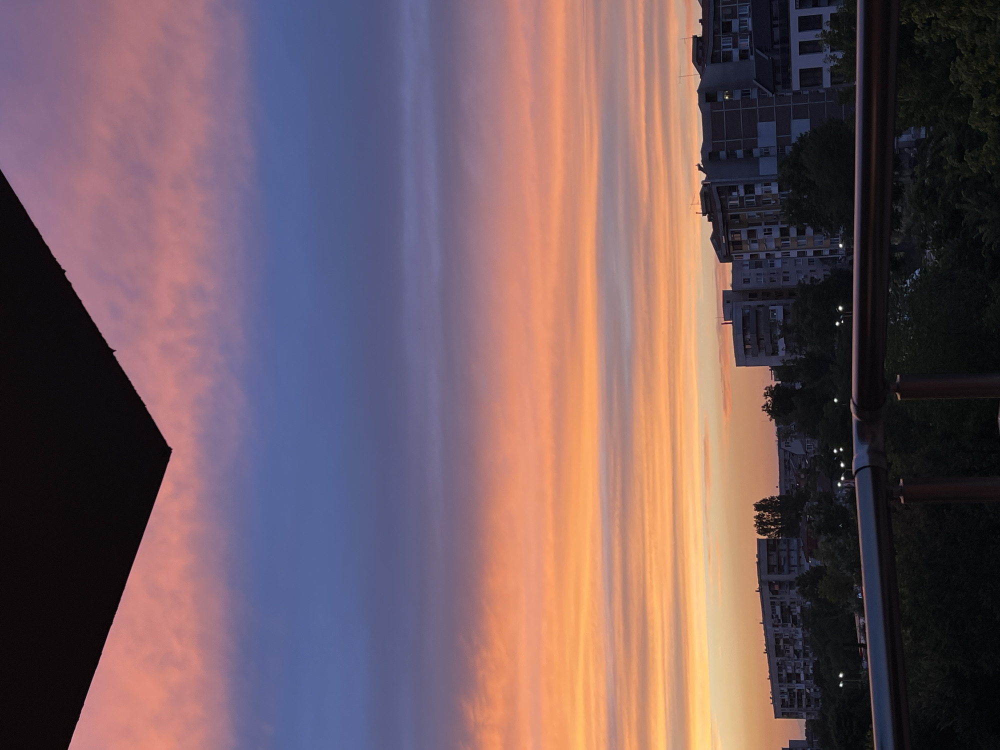 The image captures a sunset over the city of Plovdiv, Bulgaria. A dark, angular object, possibly a building overhang or awning, occupies the top left corner of the frame. Below this, the sky displays a gradient of colors, transitioning from a soft pink and orange near the horizon to a deep blue higher up. Wispy clouds are scattered across the sky, catching the hues of the setting sun. In the mid-ground, a line of apartment buildings, characteristic of Plovdiv's urban landscape, stretches horizontally across the frame. These buildings vary in height and architectural style, with visible windows and balconies. Trees and foliage are visible in front of the buildings and in the lower foreground, obscuring the ground level. A metal railing runs across the bottom of the image, suggesting the vantage point is from a balcony or elevated structure. Streetlights are visible amongst the trees, indicating it is either dusk or early evening.