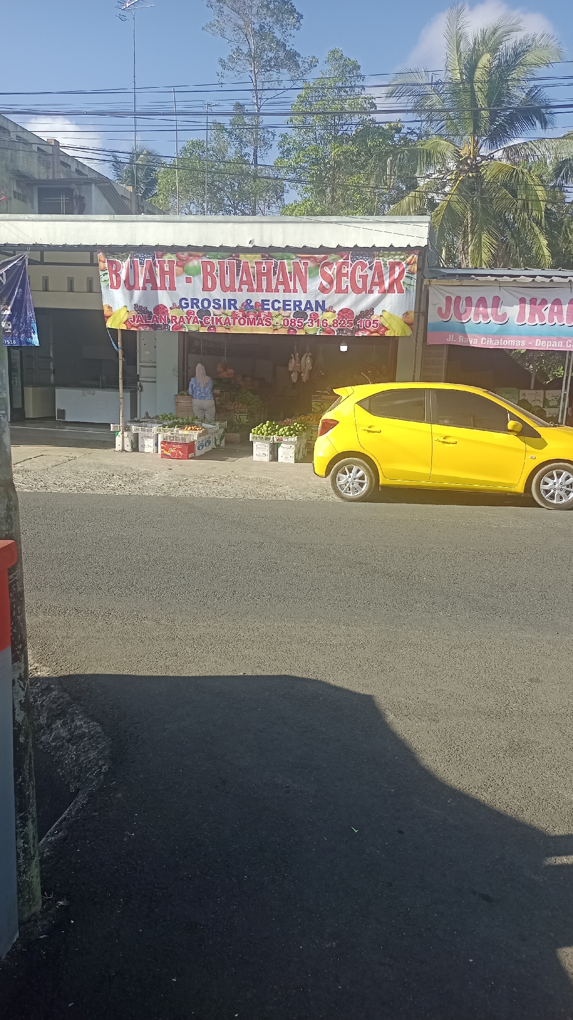 In Karyamandala, Indonesia, a yellow car is parked next to a fruit stand. The fruit stand displays an array of fresh produce, including apples and other fruits, arranged in crates. A woman in a light-colored hijab is visible behind the counter of the fruit stand. Above the stand, a banner reads "BUAH - BUAHAN SEGAR," indicating "fresh fruits." Another banner to the right advertises "JOAL IKAN," meaning "fish for sale." The scene is illuminated by sunlight, casting shadows on the asphalt road in the foreground. Trees and buildings form the backdrop.
