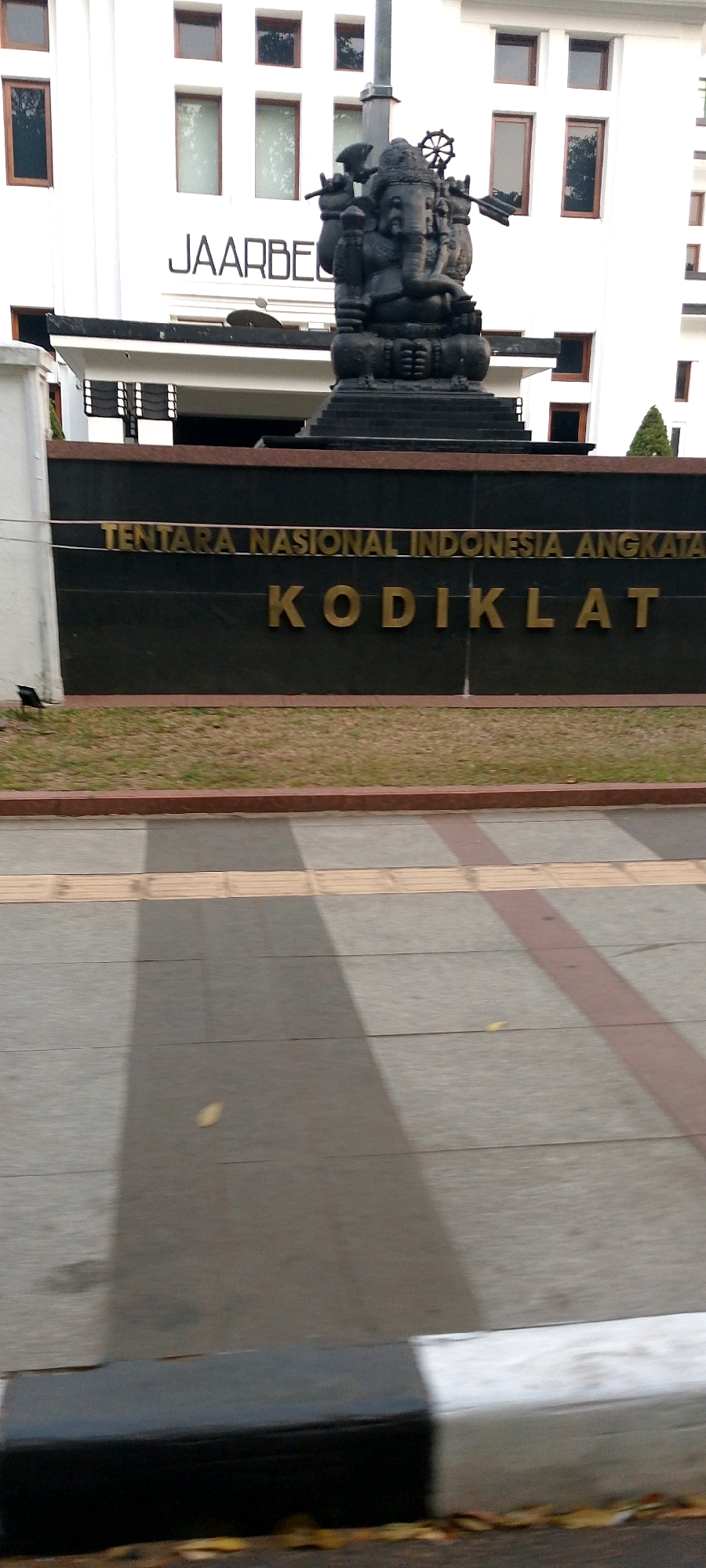 The image captures the facade of a building with a large black wall in front. The wall has golden lettering that reads "TENTARA NASIONAL INDONESIA ANGKATAN" on the top line and "KODIKLAT" on the bottom line. To the left of the wall, a portion of a white building is visible with several windows. A statue of Ganesha is prominently displayed in front of the building, positioned behind the wall. The ground in front of the wall is covered with grass and a walkway with tactile paving. The background shows the white building with multiple windows, and a street with asphalt and concrete pavement with a curb in the foreground. The image is taken outdoors, likely in a city setting. Without additional context or visible landmarks, pinpointing the exact location within Bandung City, Indonesia, is not possible based solely on this image.
