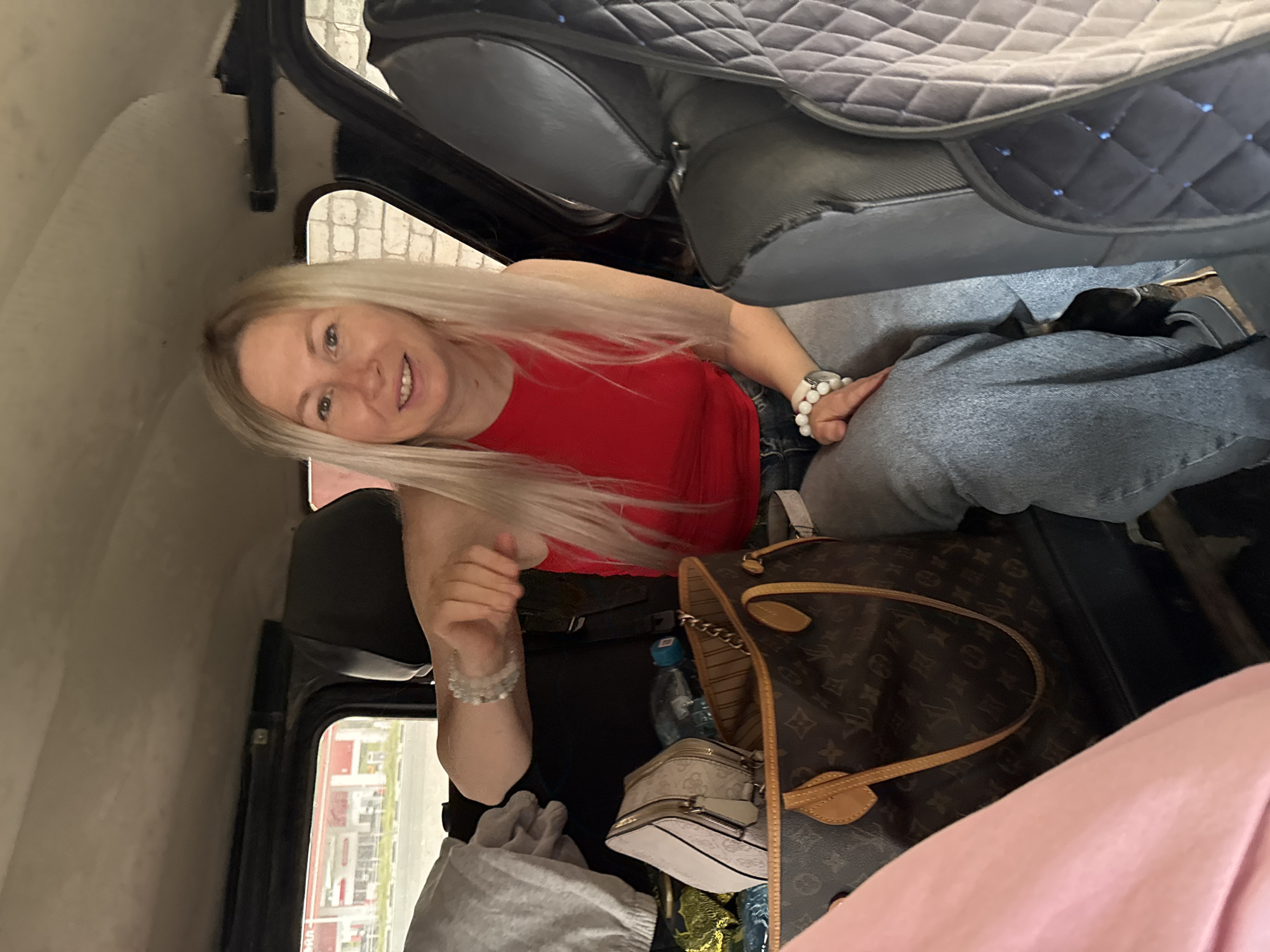 A blonde woman with long hair, wearing a red tank top and blue jeans, is sitting in the back of a vehicle, possibly a car or van. She has bracelets on her wrists. Beside her are several bags, including a large designer tote bag with a monogram pattern and a smaller white bag. A water bottle is also visible. The interior of the vehicle appears to be somewhat cluttered with personal belongings. Through the window, a building with a red roof and what appears to be a gas station or similar commercial establishment is visible in the distance, suggesting an urban or suburban environment. The overall scene depicts a person preparing for or in the midst of travel.