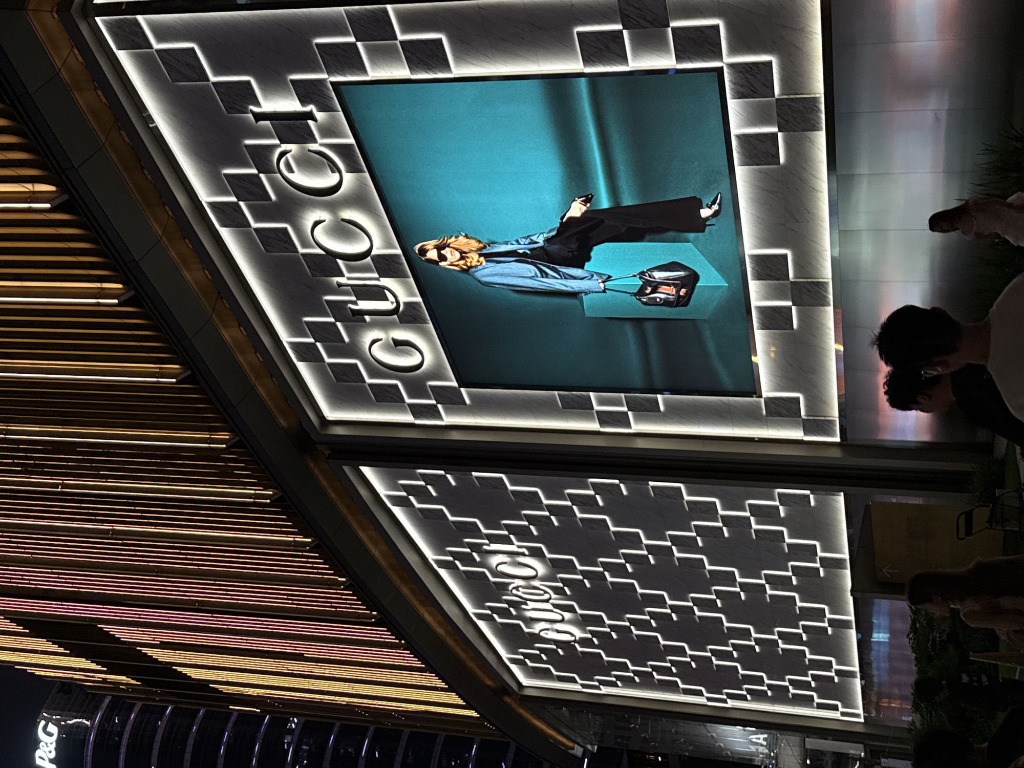 This media file captures a nighttime scene in Tianhe District, China, focusing on a stylish Gucci storefront. The primary subject is a large, illuminated display featuring a model posing elegantly against a teal backdrop. The model is dressed in a pinstripe blazer and black trousers, accessorized with sunglasses and a black handbag. The Gucci logo is prominently displayed in illuminated, stylized lettering on the facade of the building, integrated into a geometric, checkered design with white lighting.
Adjacent to the main display, another illuminated section showcases a similar geometric pattern. The building's exterior is further adorned with rows of horizontal lights, displaying warm yellow and pink hues, suggesting an urban, commercial setting at night.
In the foreground and to the right of the frame, several people are visible, partially silhouetted and out of focus, suggesting they are either patrons or passersby observing the storefront. Their presence adds a sense of scale and context to the luxury retail environment. The overall mood is sophisticated and fashionable, indicative of a high-end shopping district. No specific weather conditions are discernible. Visible text includes "GUCCI" on the storefront and a partial "P&G" logo on a building in the lower left corner.