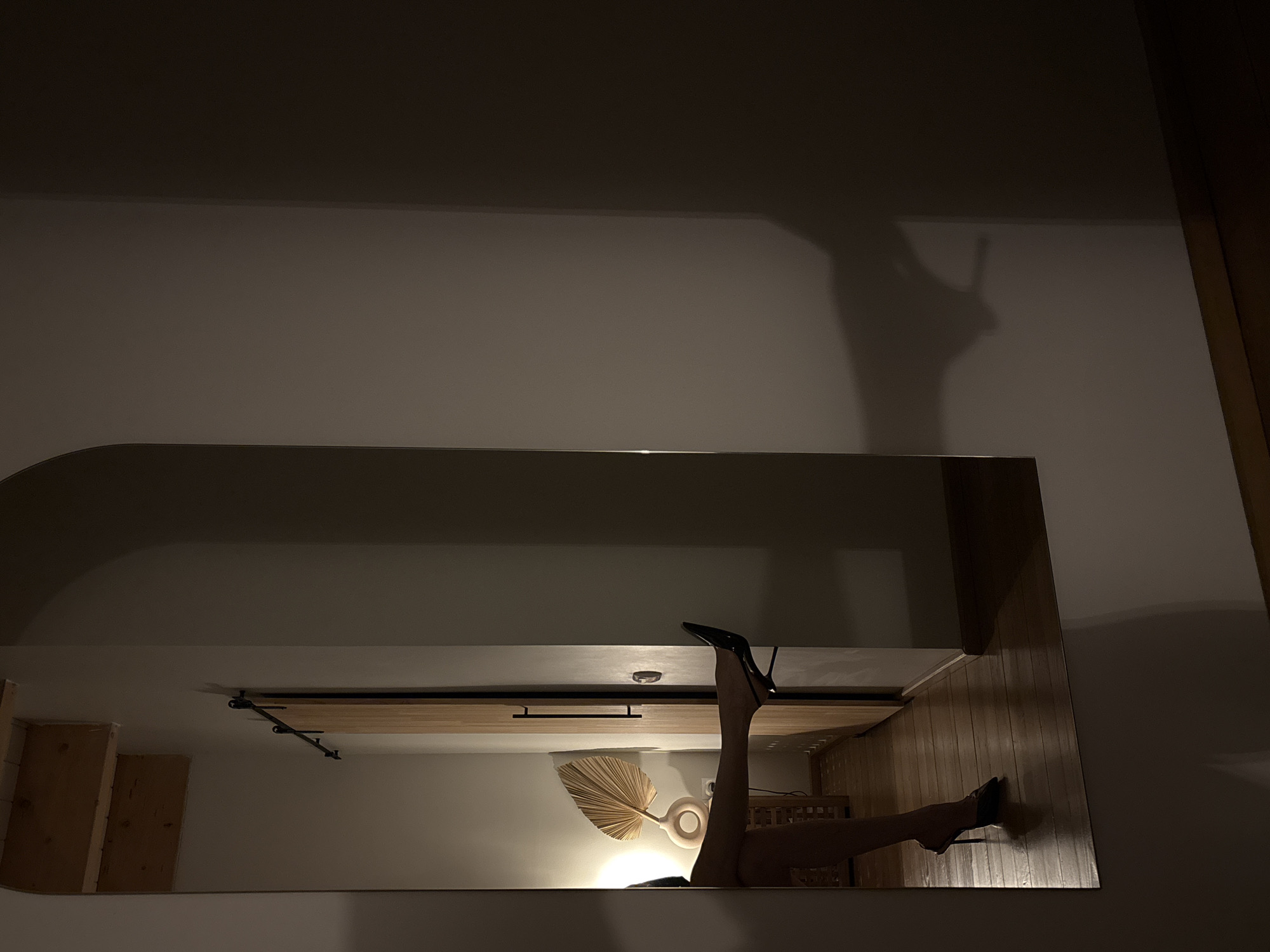 The image depicts an interior scene in Chaikovsky, Russia, characterized by low, warm artificial lighting. A large, vertical rectangular mirror occupies the left portion of the frame, reflecting a segment of a room. Within this reflection, the lower half of an individual's body is visible, specifically their legs. The left leg is extended horizontally, with the foot, clad in a black high-heeled pump, resting against a light-colored wooden sliding barn door equipped with a black vertical handle. The right leg, also wearing a black high-heeled pump, is positioned closer to the wooden floorboards. To the left of the door in the reflection, a lamp with a round base and a natural-toned, fan-shaped shade emits a soft glow from a table or stand. Exposed wooden ceiling beams are visible in the upper part of the reflected area. The right side of the image displays a plain, light-colored wall, featuring a distinct shadow of a high-heeled shoe cast upon it. An arched architectural recess is partially visible in the upper right corner of this wall. The entire visible floor, both reflected and direct, consists of wooden planks.