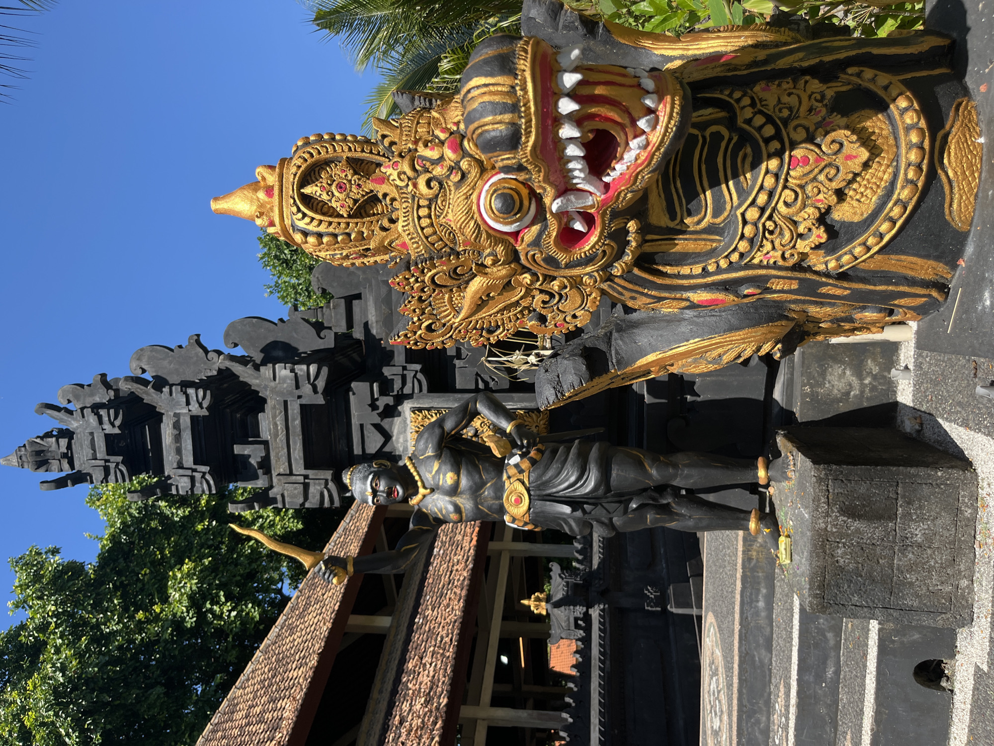 The image depicts a scene in Kuta Selatan, Indonesia, featuring traditional Balinese temple statues. In the foreground, to the right, is a large, ornately decorated statue of a dragon's head, predominantly black with intricate gold and red detailing. Its mouth is open, revealing sharp white teeth and a red interior. To the left of the dragon statue stands a dark-skinned humanoid statue with a muscular physique, holding a golden dagger aloft in its right hand. This figure wears a golden necklace, a golden belt with a red circular emblem, and a dark wrap around its waist and legs. Behind these statues, a traditional Balinese temple structure with a tiered, dark gray roof and black carved details is visible. Lush green trees and a clear blue sky form the backdrop. The statues are positioned on stone platforms and steps, with the ground covered in light-colored gravel and decorative white stones.