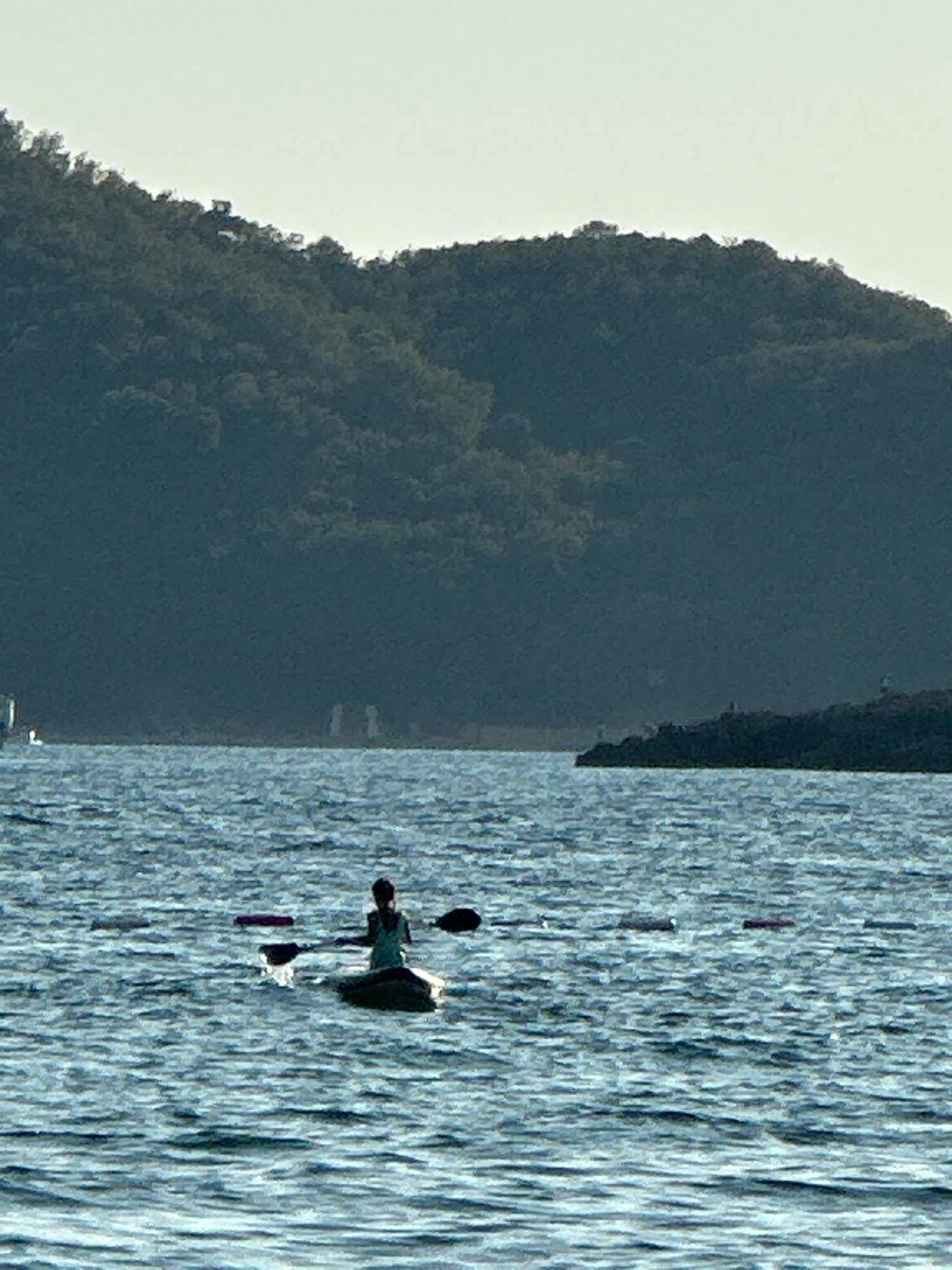 The image depicts a serene scene on the water, likely in the vicinity of Boreti, Montenegro, with a backdrop of a tree-covered hill. In the foreground, a person is seated in a kayak, paddling across the calm blue water. The kayak appears to be dark in color. The person is silhouetted against the water, and their posture suggests they are actively engaged in kayaking. The water is rippled, indicating gentle movement. In the distance, a dark, rocky shoreline is visible, with trees lining the slopes of the hill. The sky above is a pale, hazy blue, suggesting either early morning or late afternoon light. There are some distant objects that appear to be buoys or markers on the water, as well as some structures or buildings visible on the hillside in the far distance, though they are indistinct.