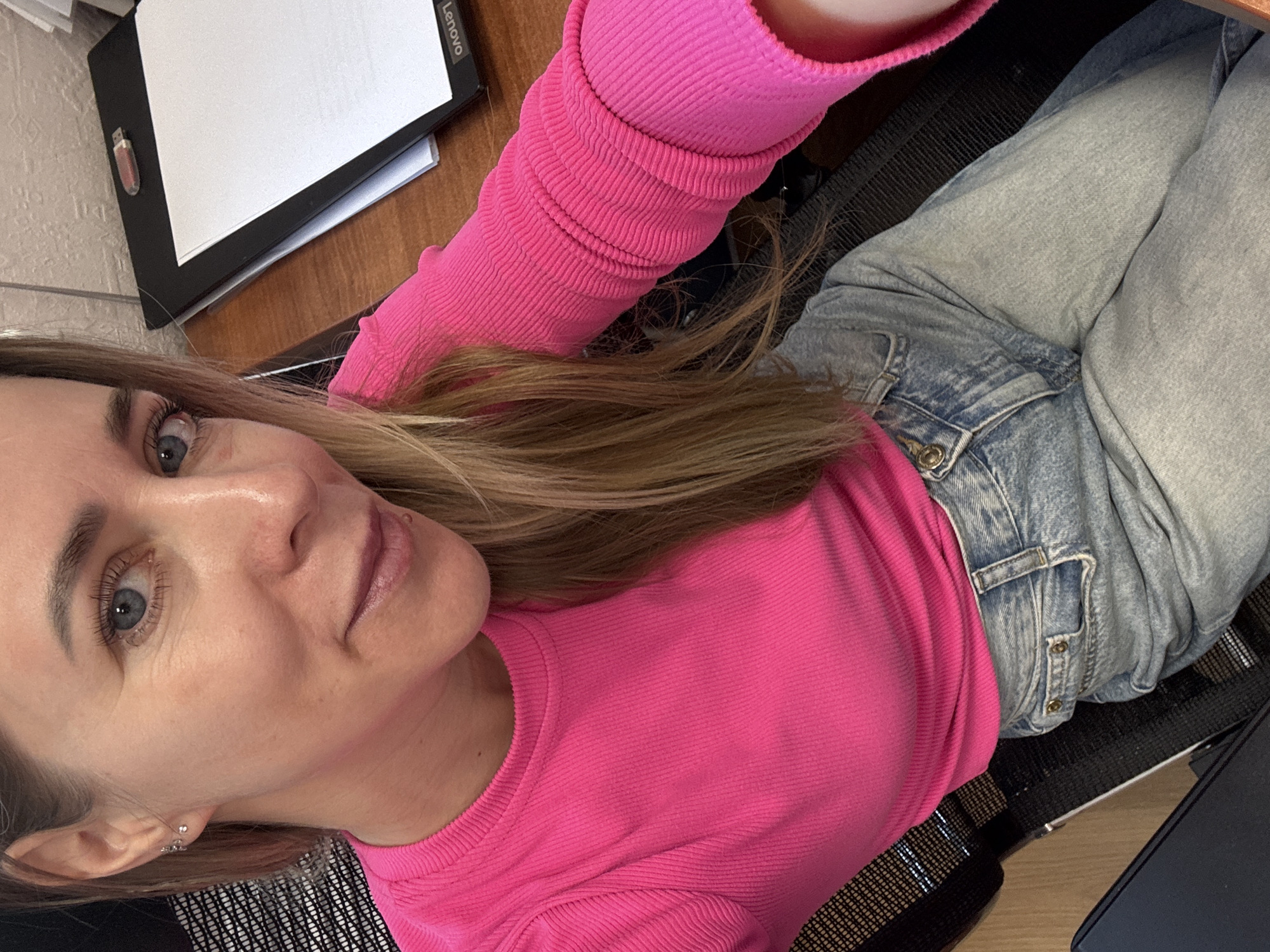 A woman is seated at a desk, wearing a bright pink long-sleeved shirt and light wash jeans. She is angled towards the camera, looking directly at it. Her hair is blonde and slightly parted, falling around her face. On the desk to her left is a black folder with papers inside, and a USB drive is attached to the folder. The location is Chaikovsky, Russia, based on visual analysis of the surrounding environment.