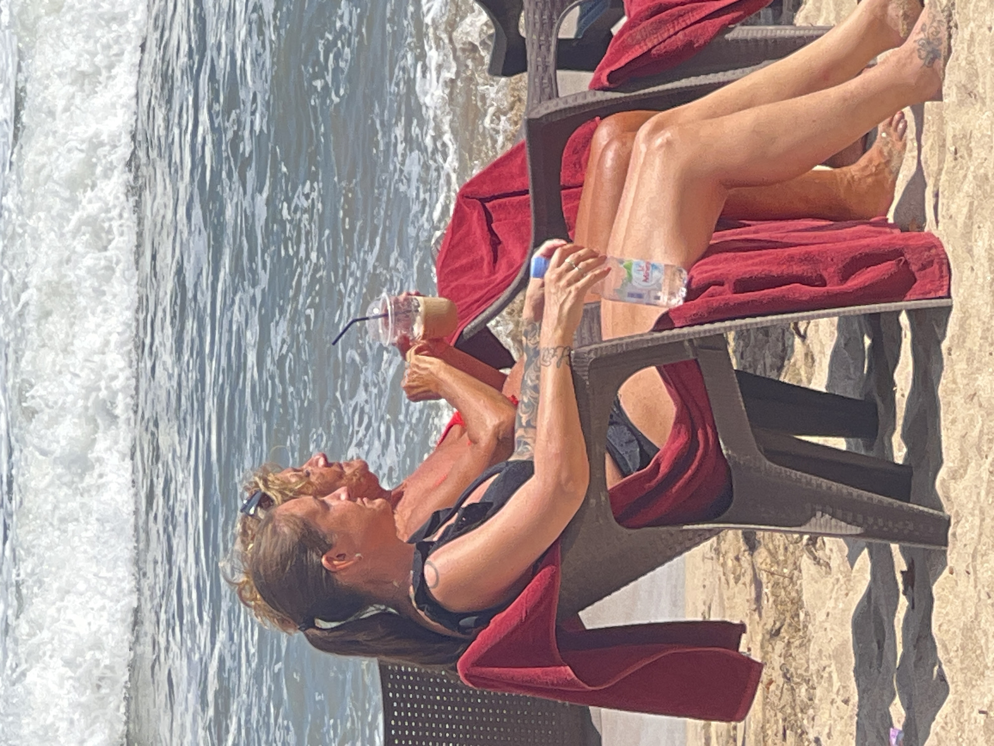 Two women are seated on beach chairs facing the ocean in Kuta, Indonesia. The woman in the foreground is wearing a black bikini and has a ponytail and tattoos on her arm. She is holding a bottle of water. The woman behind her is wearing a red bikini and has curly blonde hair. Both women are looking out at the ocean. The beach is sandy, and there are red towels draped over the chairs. The ocean has waves, and the sky is bright and sunny.