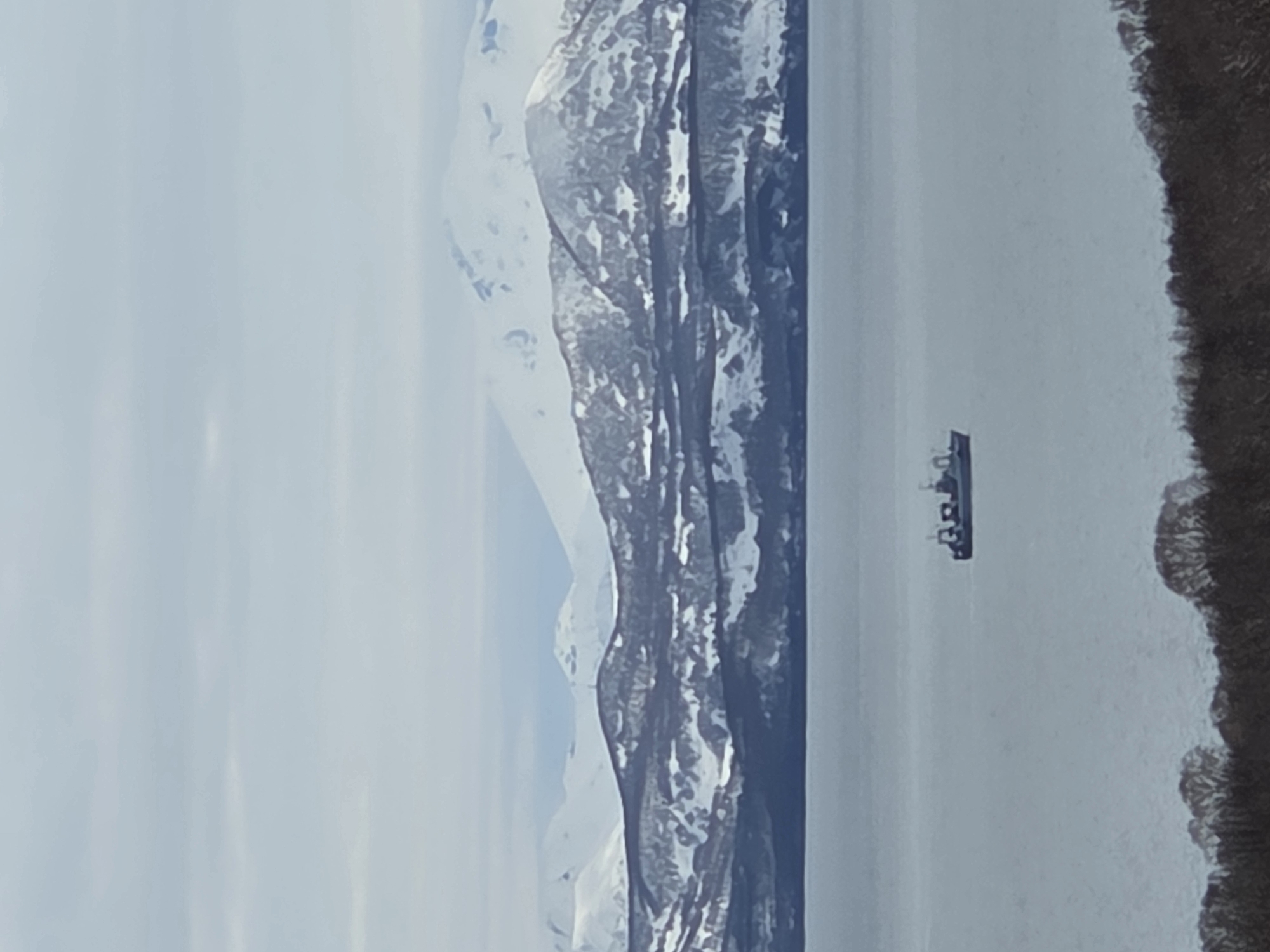 The scene depicts a body of water, possibly a lake or a fjord, with a vessel navigating its surface. The vessel appears to be a small ship or a large boat, possibly a ferry. Snow-capped mountains line the left side of the water body, while a landmass covered in what looks like trees or low-lying vegetation is seen on the right. The sky is overcast. The location in the city is Unknown, Unknown.