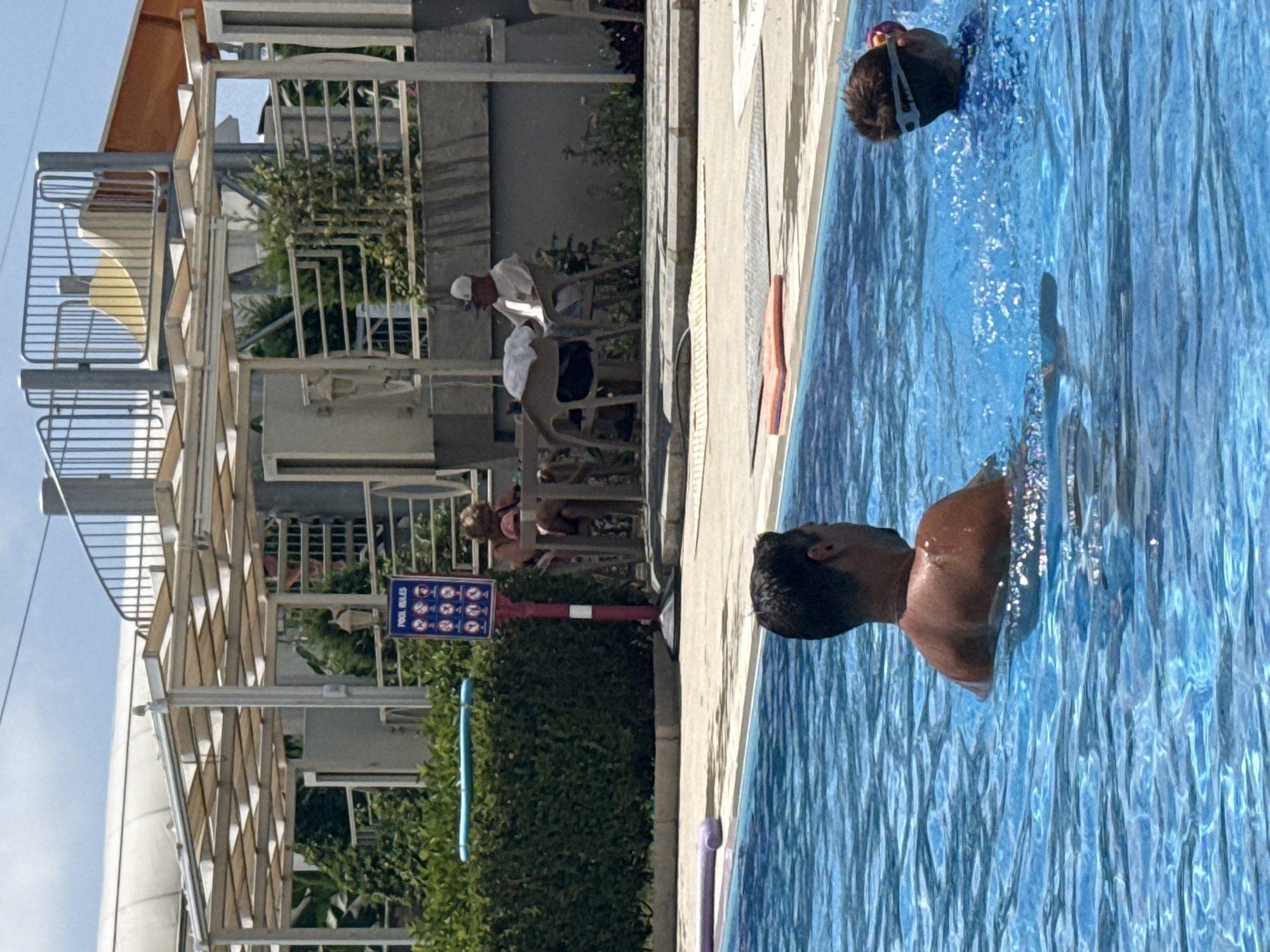 The image depicts a sunny day at an outdoor swimming pool, likely in Alanya, Turkey, given the presence of a water slide structure and lush greenery characteristic of the region. In the foreground, two individuals are partially submerged in the bright blue water. A man with dark hair is facing away from the camera, his back and shoulders visible as he floats in the pool. To his right, a child wearing goggles is swimming, their head and upper body visible above the water's surface. The water is disturbed, suggesting movement.
In the mid-ground, a trimmed green hedge forms a border behind the pool. A red and white pole stands near the hedge, displaying a blue sign with multiple icons labeled "POOL RULES." Behind the hedge, under a white canopy structure, a woman and a man are seated at a table. The woman is wearing a pink top and appears to be looking down, while the man, wearing a white cap, is facing to the left and looking forward. Two chairs are visible around the table, one of which is occupied by the man. Another empty chair is to his left.
The background reveals elements of a resort or water park. A silver metal railing with vertical bars runs along a raised structure, leading to what appears to be a portion of a yellow and orange water slide. The overall environment is bright and appears to be well-maintained, with a clear sky above.
