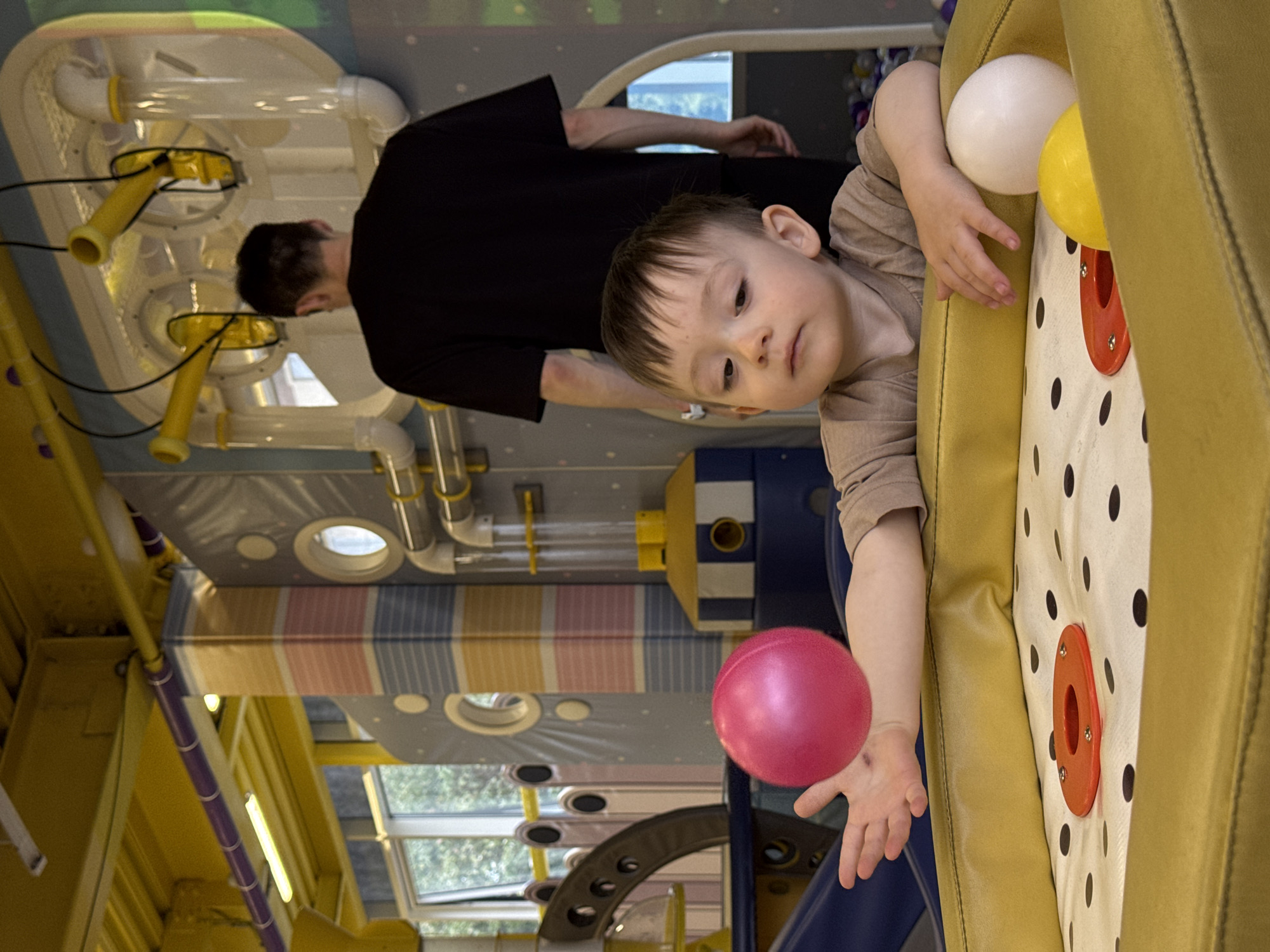 A young child plays at an indoor play area. The child, wearing a light brown shirt, reaches for a pink ball. Beside the child are white and yellow balls. The child is adjacent to a yellow padded structure with black polka dots and orange circular insets. A man in a black shirt stands in the background, near a series of yellow and white pipes and tubes. The environment includes colorful striped walls, a window showing trees outside, and other play structures. Based on the metadata, the photograph was taken in Sochi, Russia.