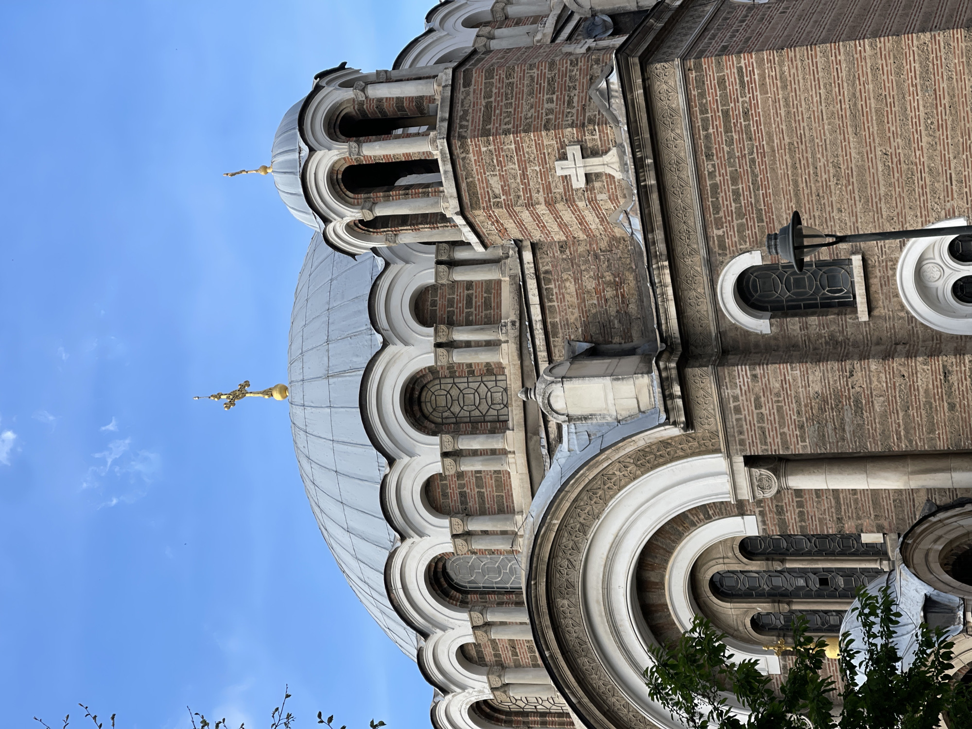 The image shows a portion of the Saint Alexander Nevsky Cathedral in Sofia, Bulgaria, viewed from a low angle. The architectural details of the cathedral are prominent, including its dome covered in gray-painted metal panels, arched windows and colonnades, and brick walls. The brickwork has alternating light and dark bricks arranged in vertical rows. There are several golden crosses atop the domes. The sky is visible in the background, with a clear blue color. Parts of trees are visible in the foreground. A street light is affixed to the building with geometric windows in the background. A cross is visible on the upper building.