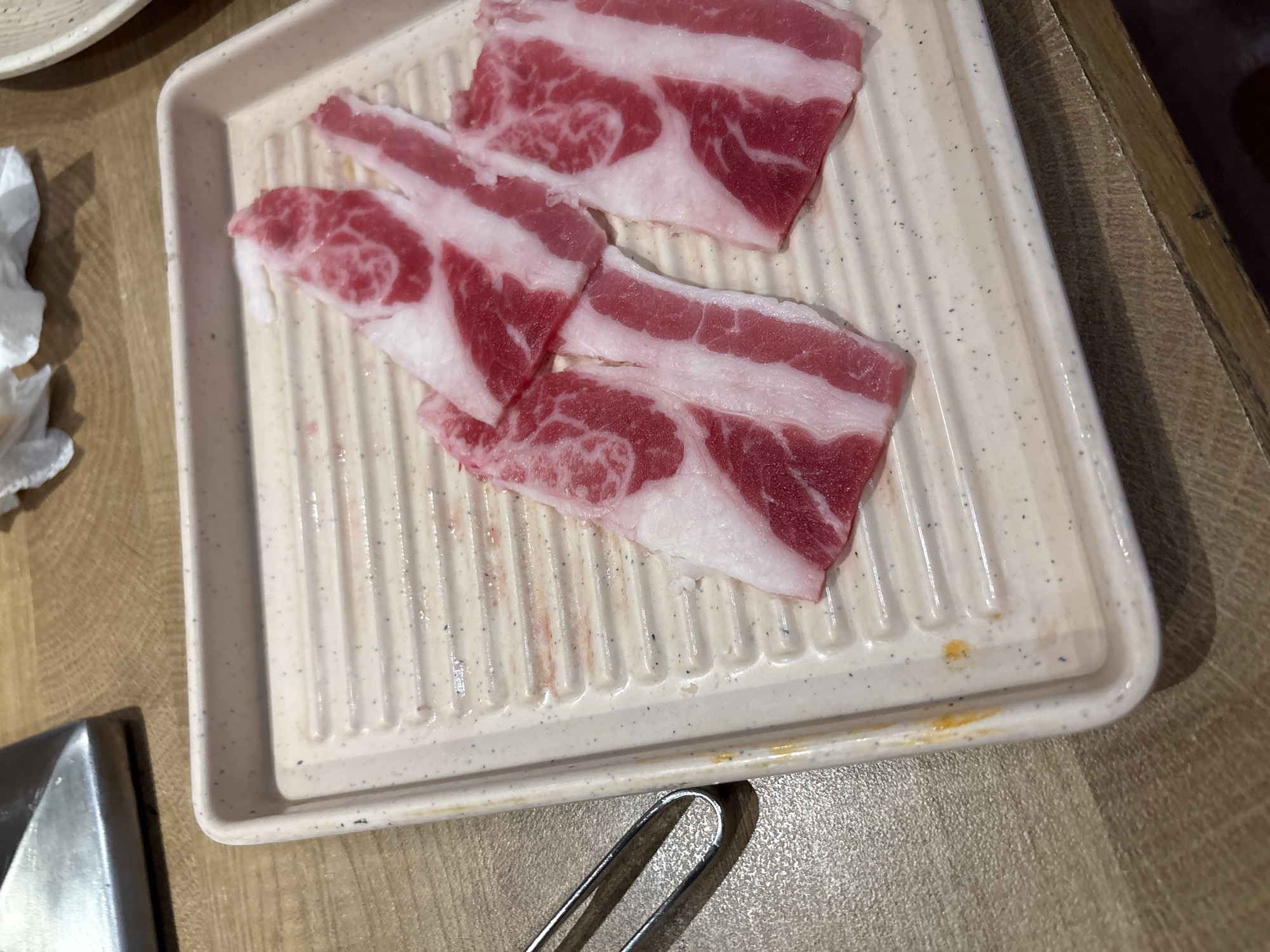 A plate of raw, thinly sliced marbled beef is displayed on a white, ribbed rectangular tray. The beef, with its distinct red and white striations, is arranged in the upper right quadrant of the tray. The tray sits on a wooden table, with a portion of a metal utensil, likely tongs, visible on the left side of the frame. A stainless steel appliance, possibly part of a cooking apparatus, is partially visible in the top left corner. The background is out of focus, suggesting an indoor setting, likely a restaurant. The image does not contain information that would allow for the determination of its location in Cirebon, Indonesia.