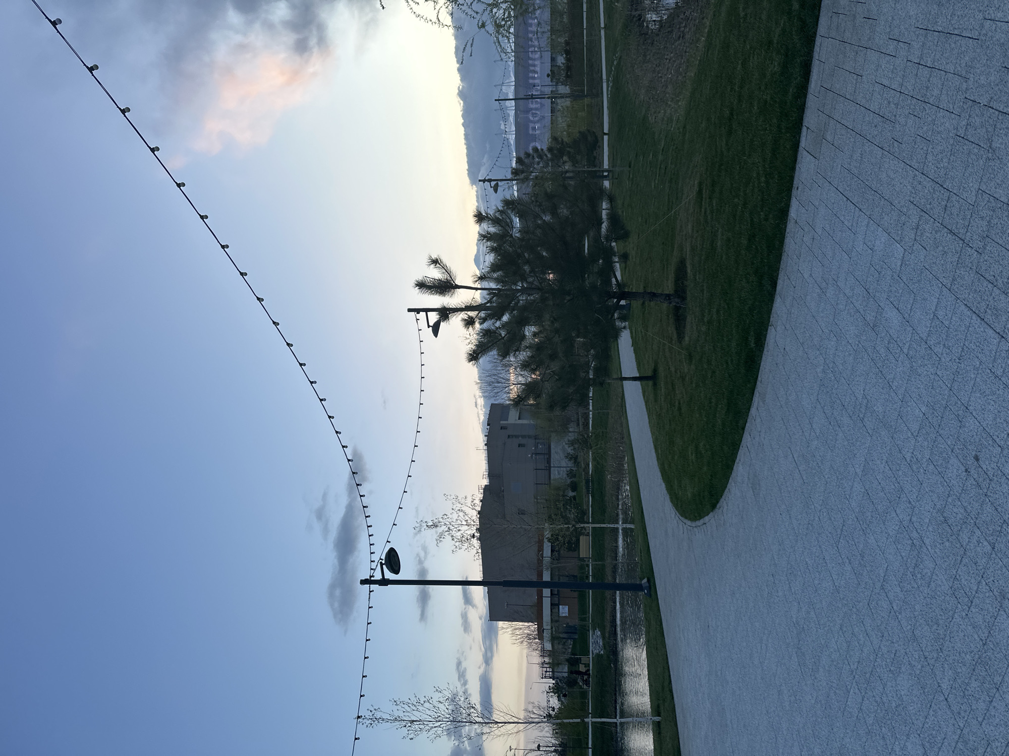 A park in Rostov-on-Don, Russia, features a curved, paved pathway bordered by grass. In the background, structures are partially obscured by trees. String lights are suspended above the park. Lighting fixtures stand along the path. The sky is cloudy with a hint of sunset.